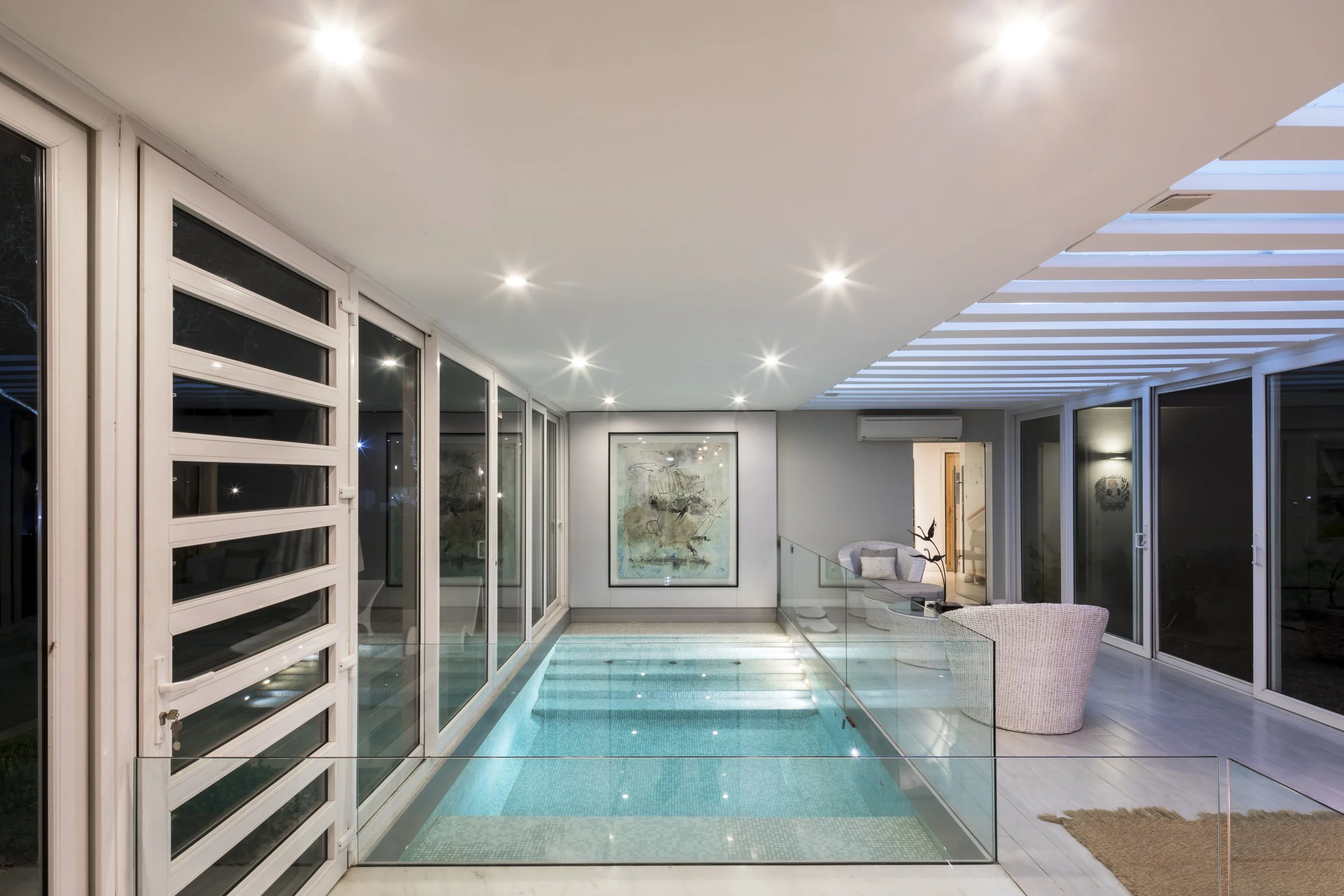 Sala de estar con piscina interior, ventanas grandes, iluminación moderna y muebles de fibra y tela en tonos neutros.