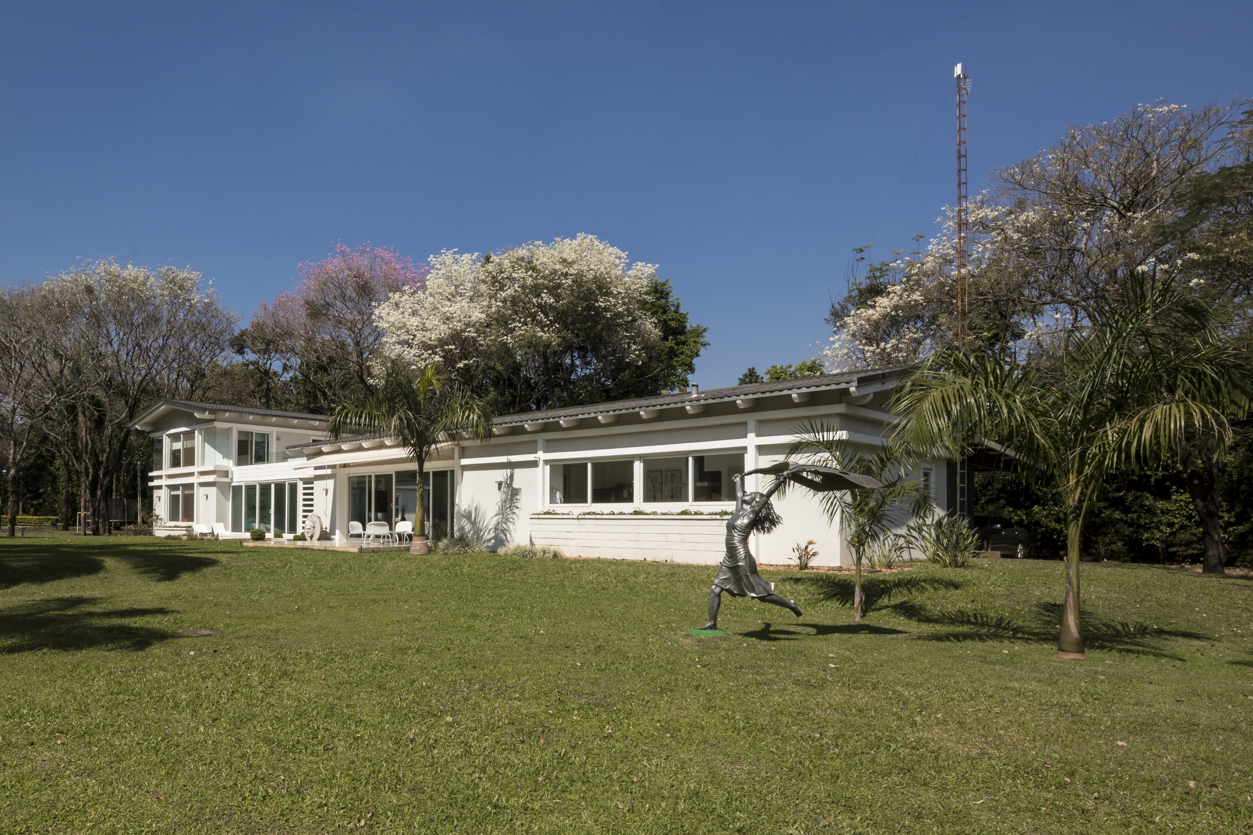 Casa blanca de un piso con ventanas largas, rodeada de árboles, con una escultura de una persona corriendo en el césped y palmeras cercanas, bajo un cielo azul claro.