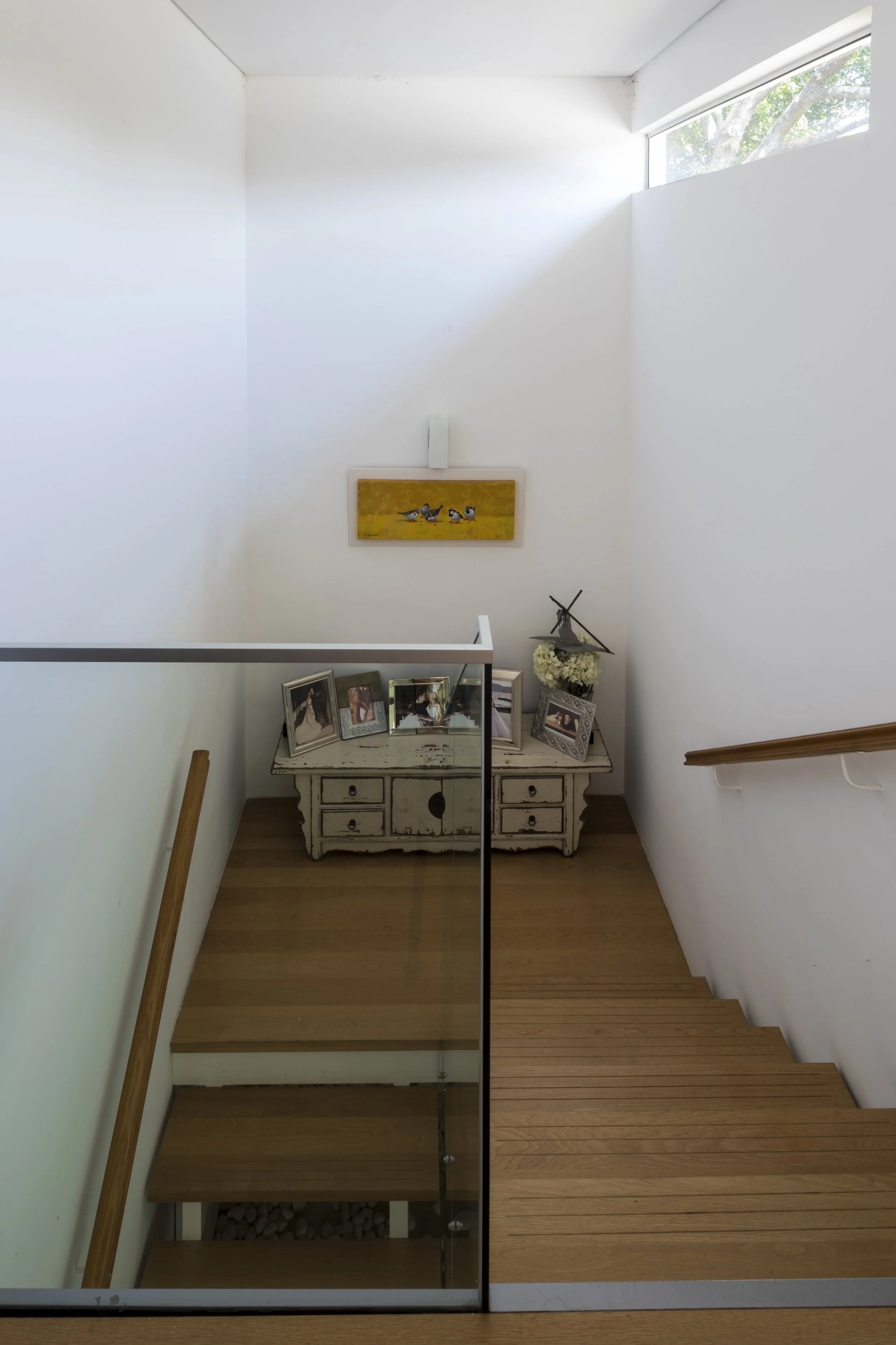 Una escalera de madera con barandilla de vidrio y una repisa con fotos familiares y decoraciones, en una casa moderna con paredes blancas y una ventana larga que deja entrar luz natural.