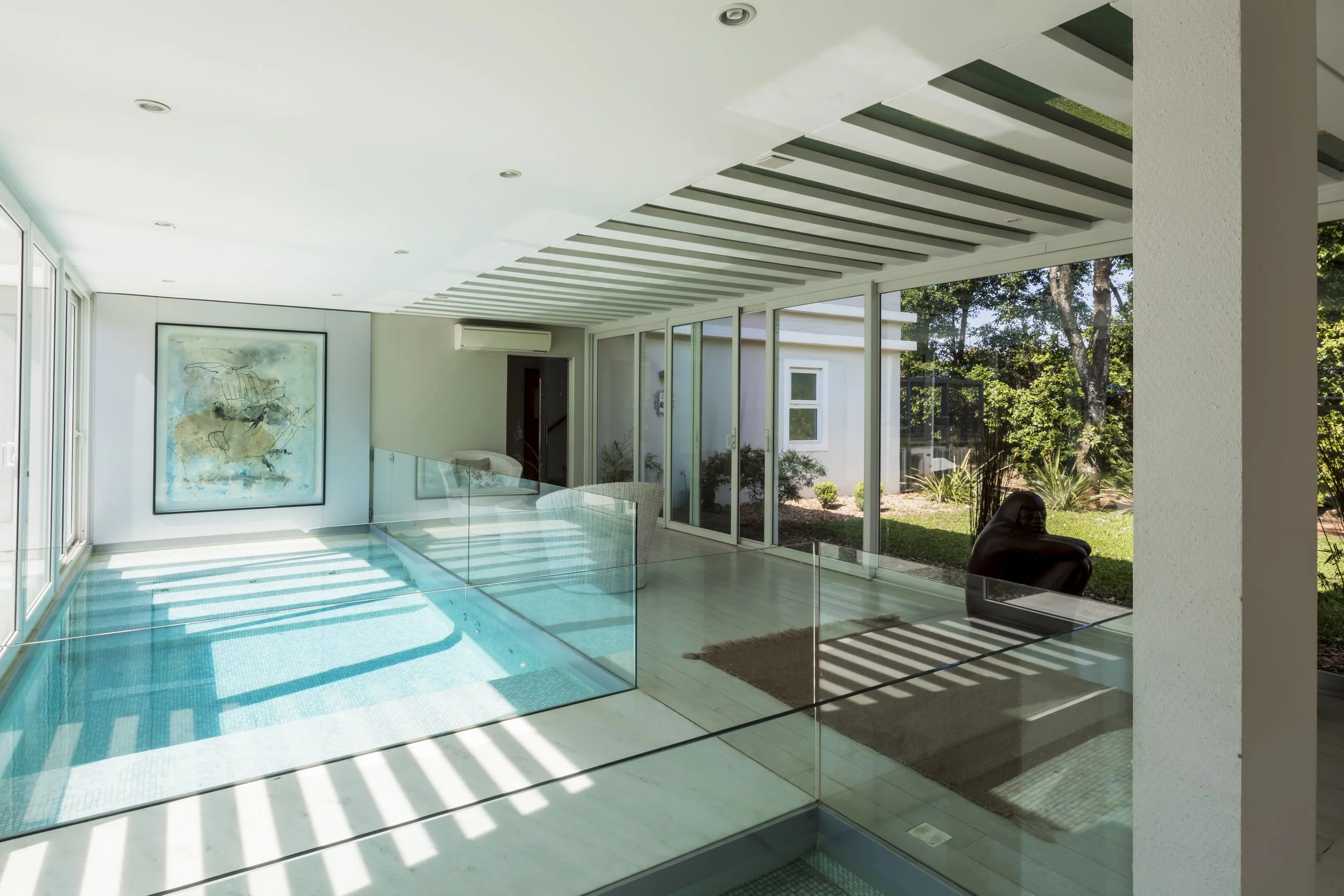 Interior de una sala moderna con paredes blancas, ventanas grandes, y una piscina con barandilla de cristal, con vista hacia un jardín exterior con árboles y plantas.