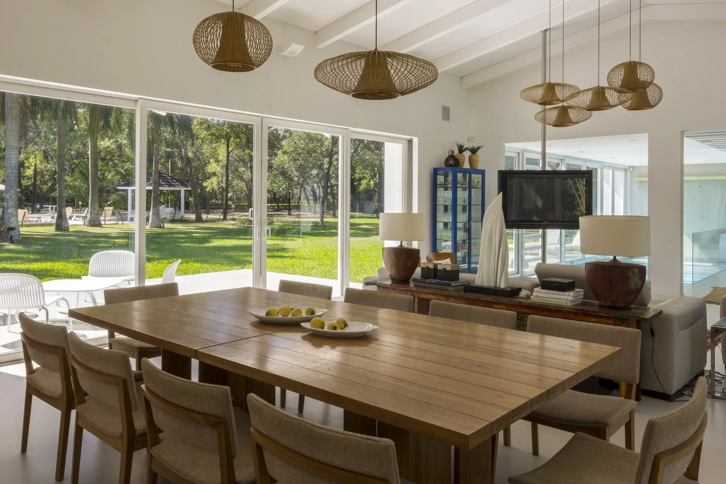Sala de comedor moderna con mesa de madera y ocho sillas, en un espacio luminoso con ventanales grandes que muestran un jardín exterior con árboles y césped.