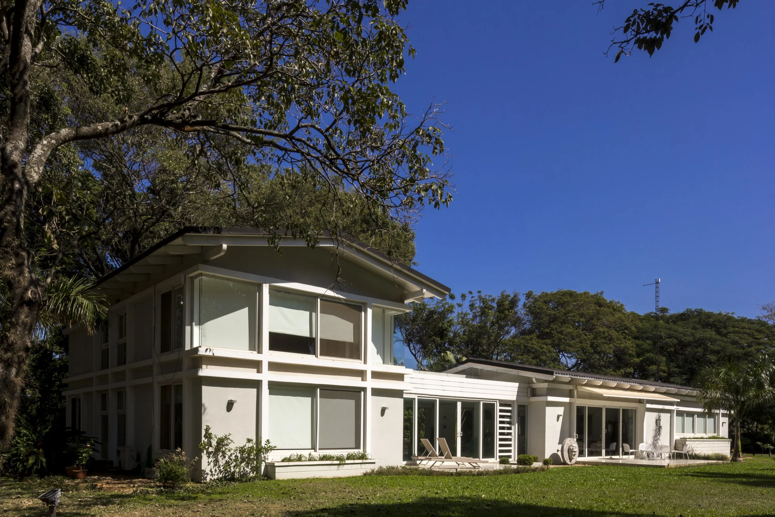 Casa moderna con amplias ventanas y jardín en un día soleado, rodeada de árboles y césped verde.