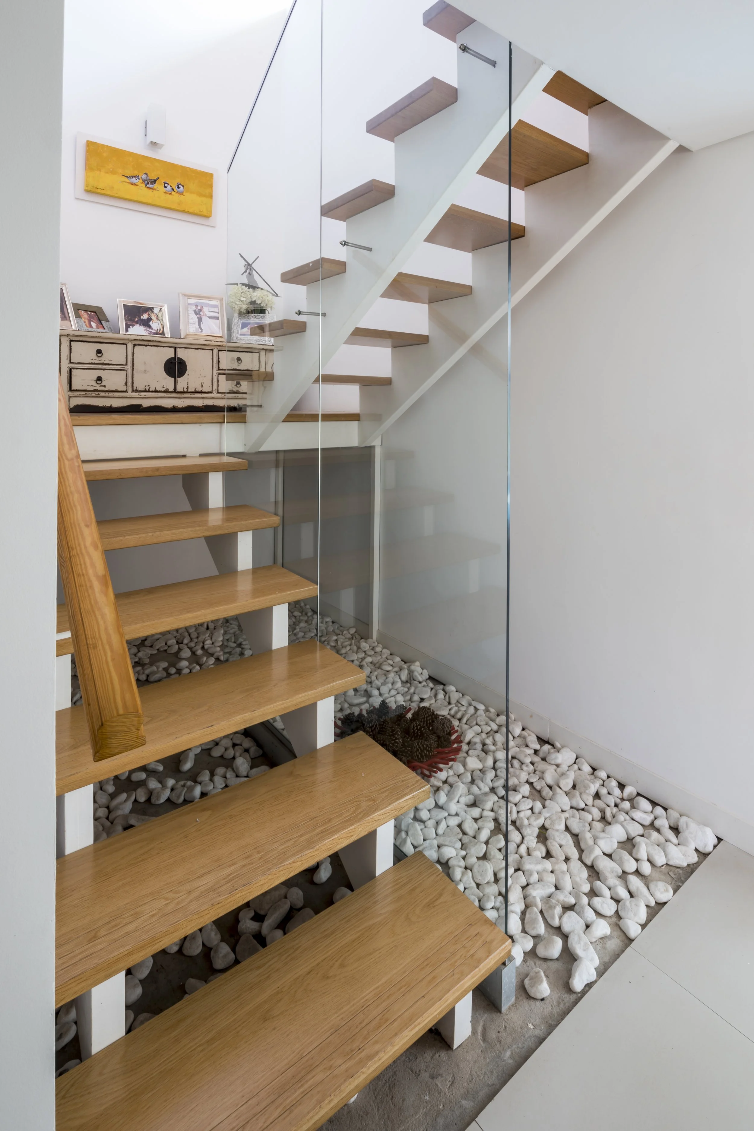 Escalera moderna con peldaños de madera y barandilla de vidrio en interior con decoración de cuadros, fotos y objetos en la repisa, y piso de piedra blanca en la base.