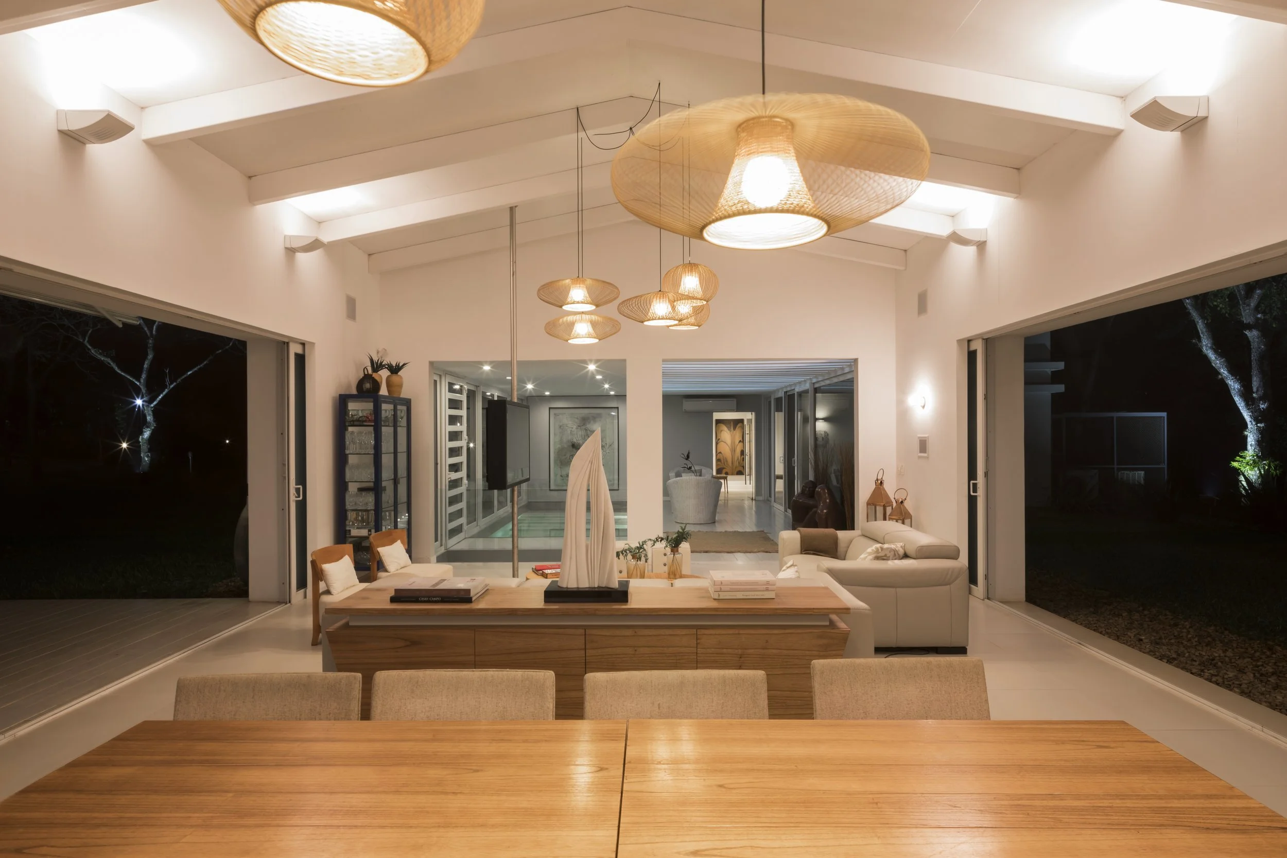 Interior de una sala moderna con iluminación cálida, mesas de madera, sillones y decoración minimalista, con ventanas grandes que muestran un ambiente exterior oscuro.