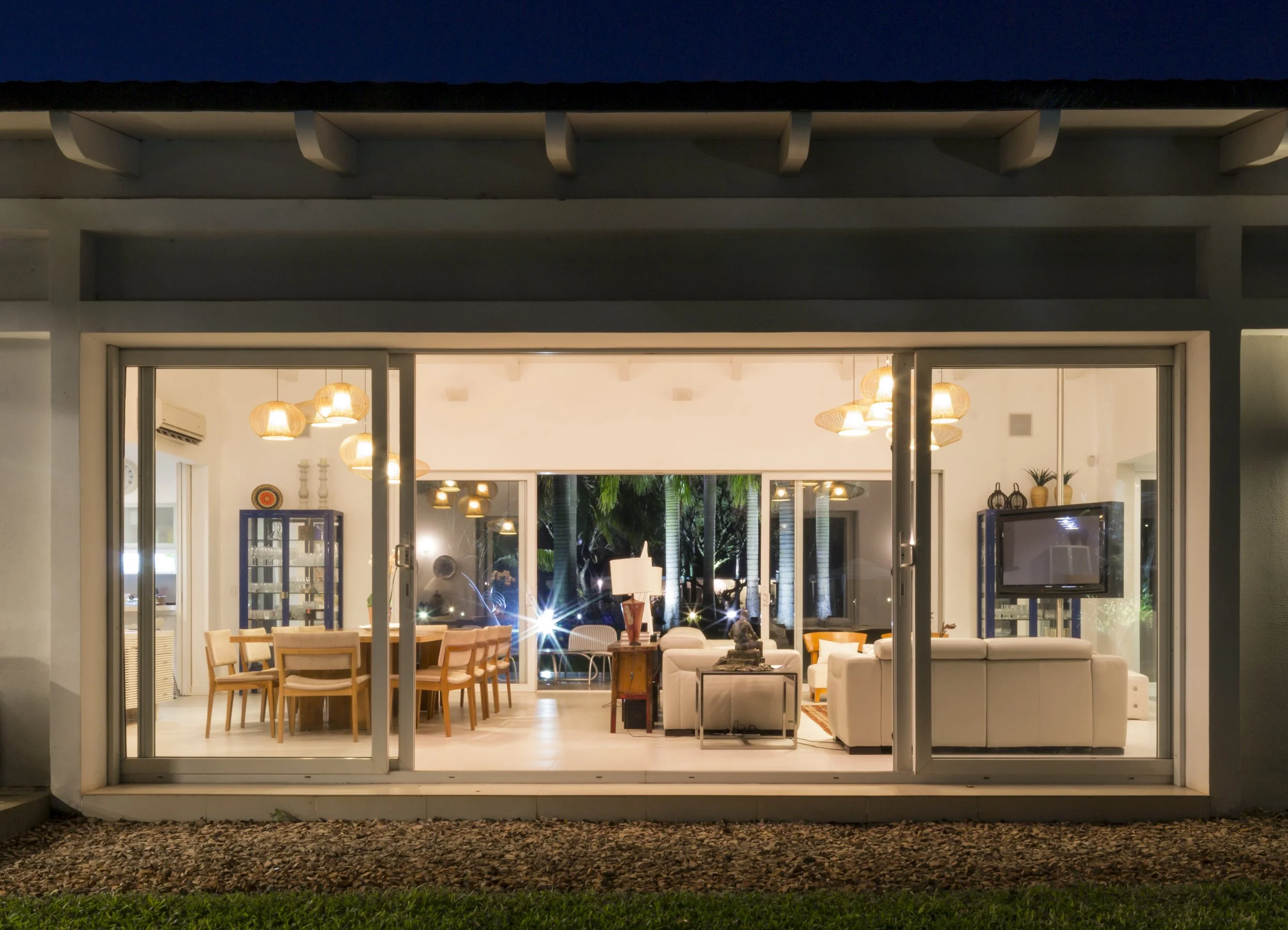 Vista de una sala de estar y comedor iluminada desde afuera, con muebles blancos, pared de ventanas y decoración moderna en la noche.