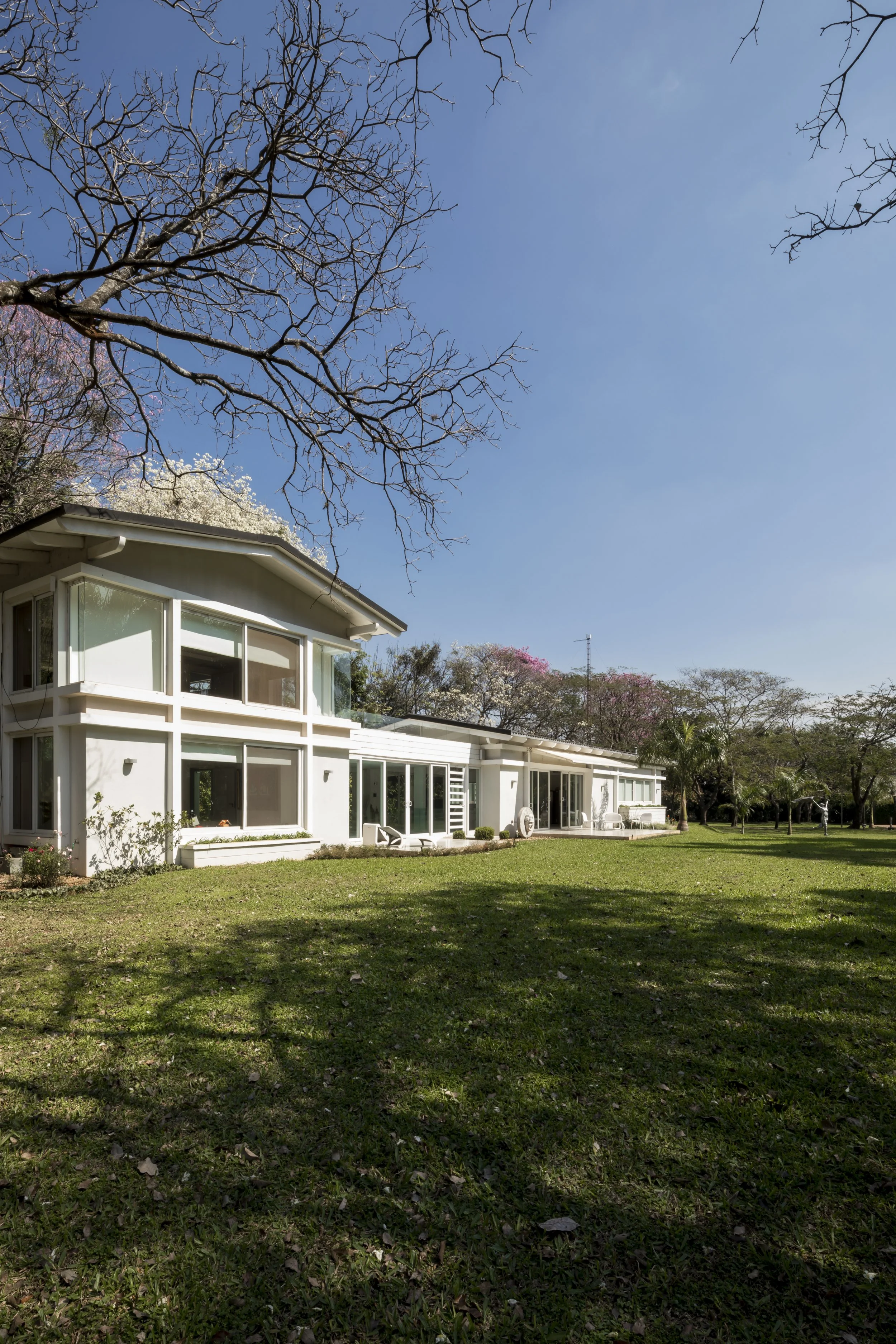 Casa moderna blanca con amplios ventanales, rodeada de césped y árboles, bajo un cielo despejado.