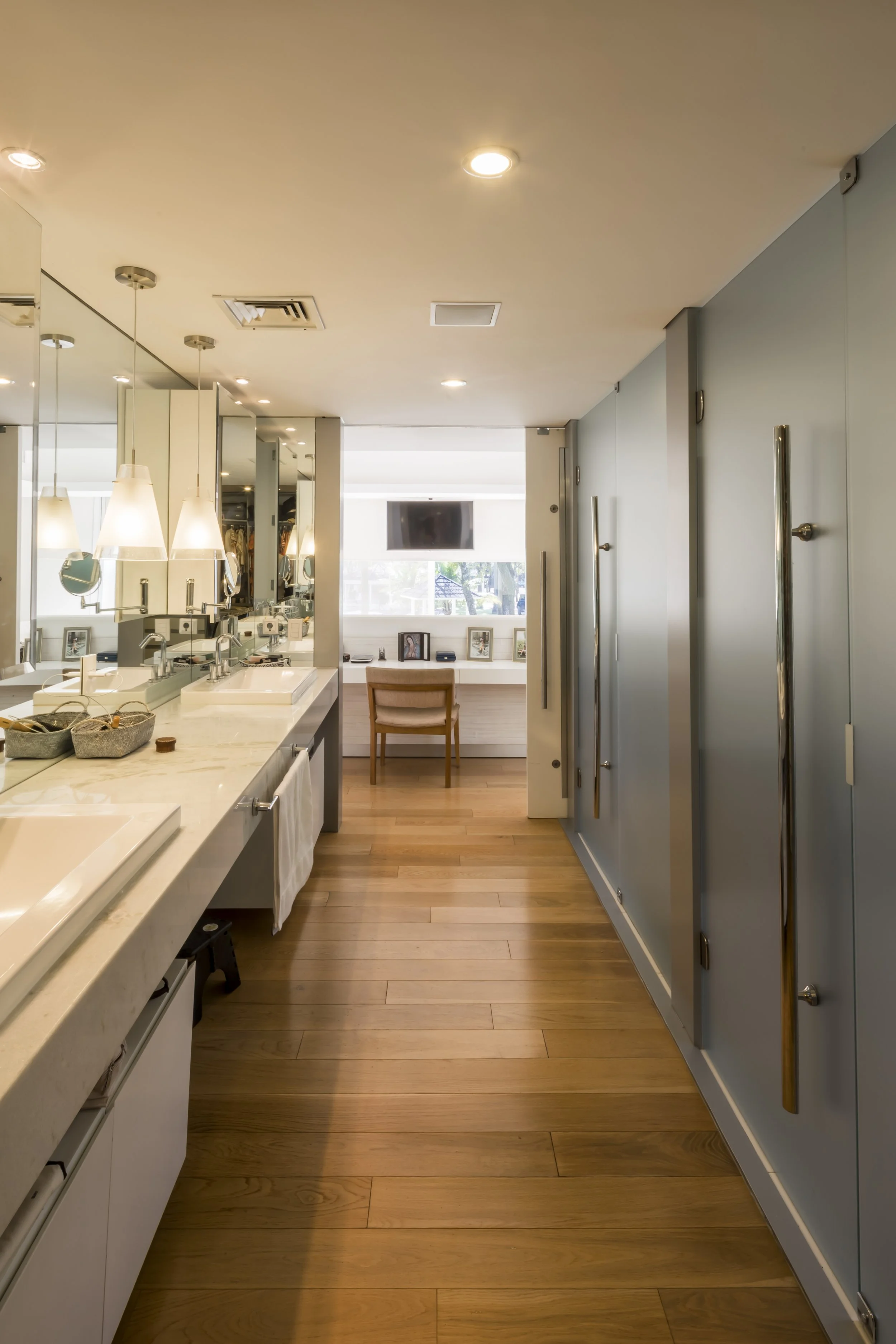 Vista interior de un baño moderno con pared de espejos, lavabos con encimeras de mármol y puertas de armario con barras metálicas, con madera en el piso y una ventana al fondo.