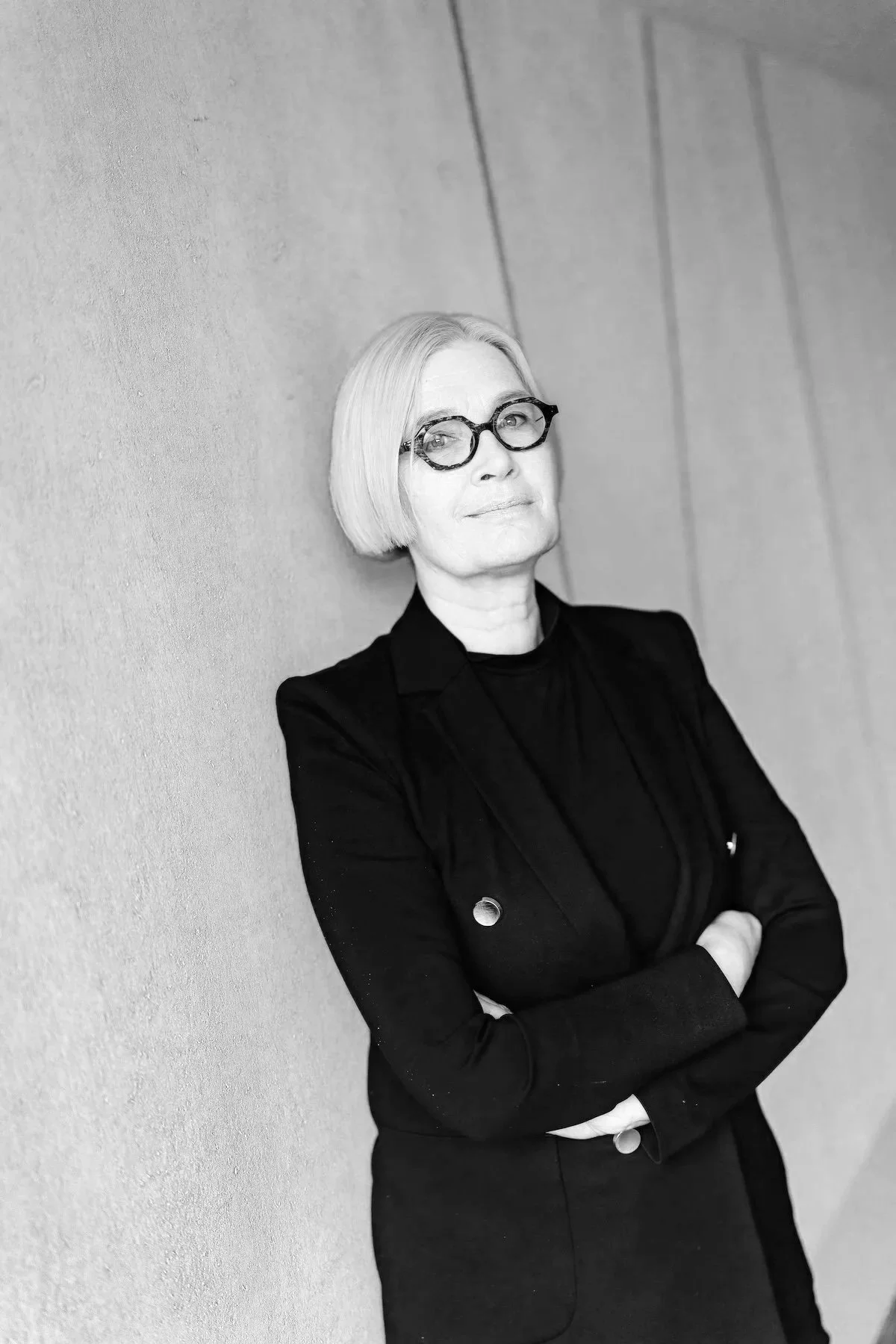 Valkoinen nainen silmälaseilla ja mustassa puvussa seisoo seinää vasten ja katsoo kameraan.