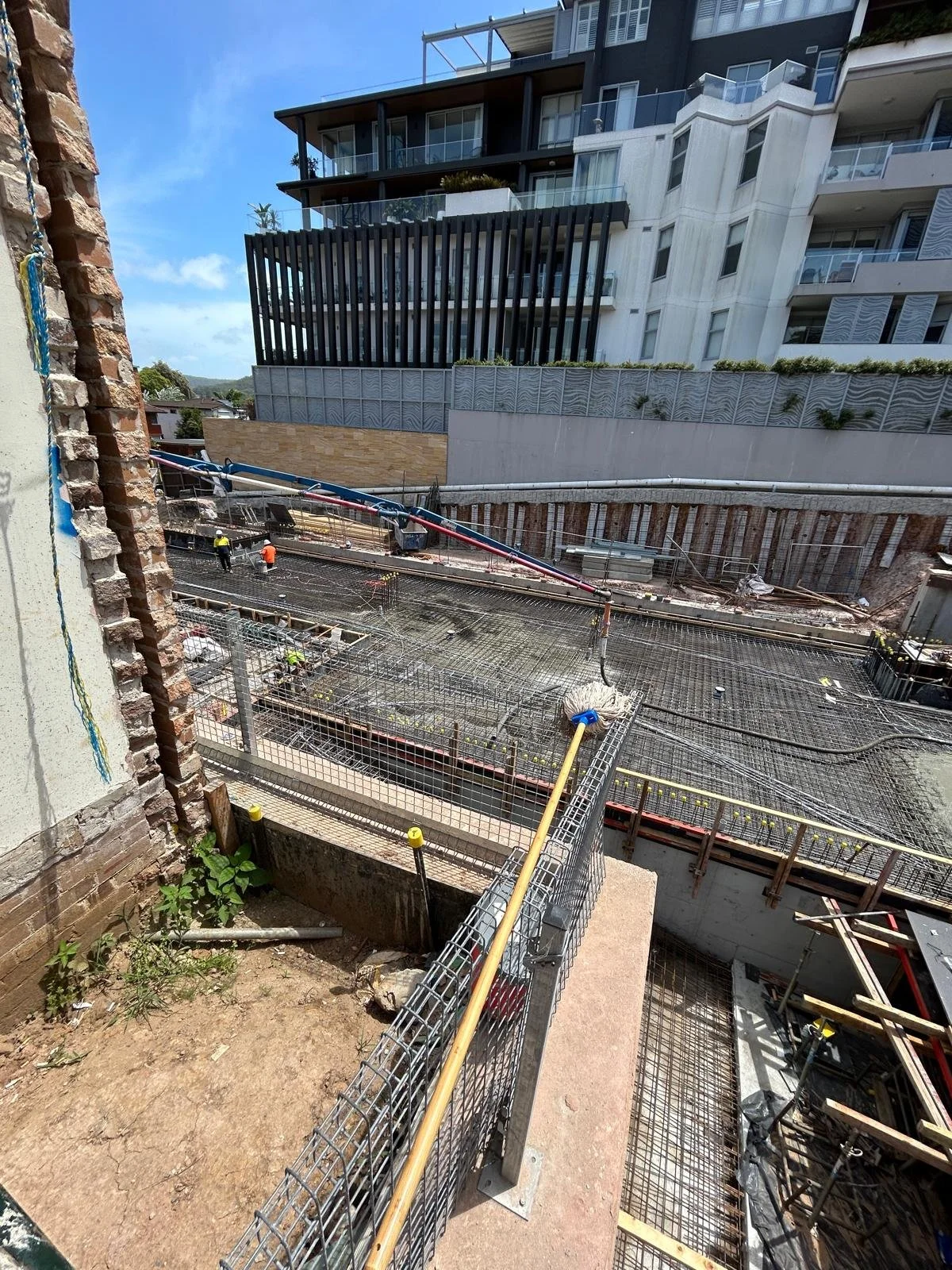 Construction site with workers and construction materials in front of modern multi-story building, with a clear blue sky.