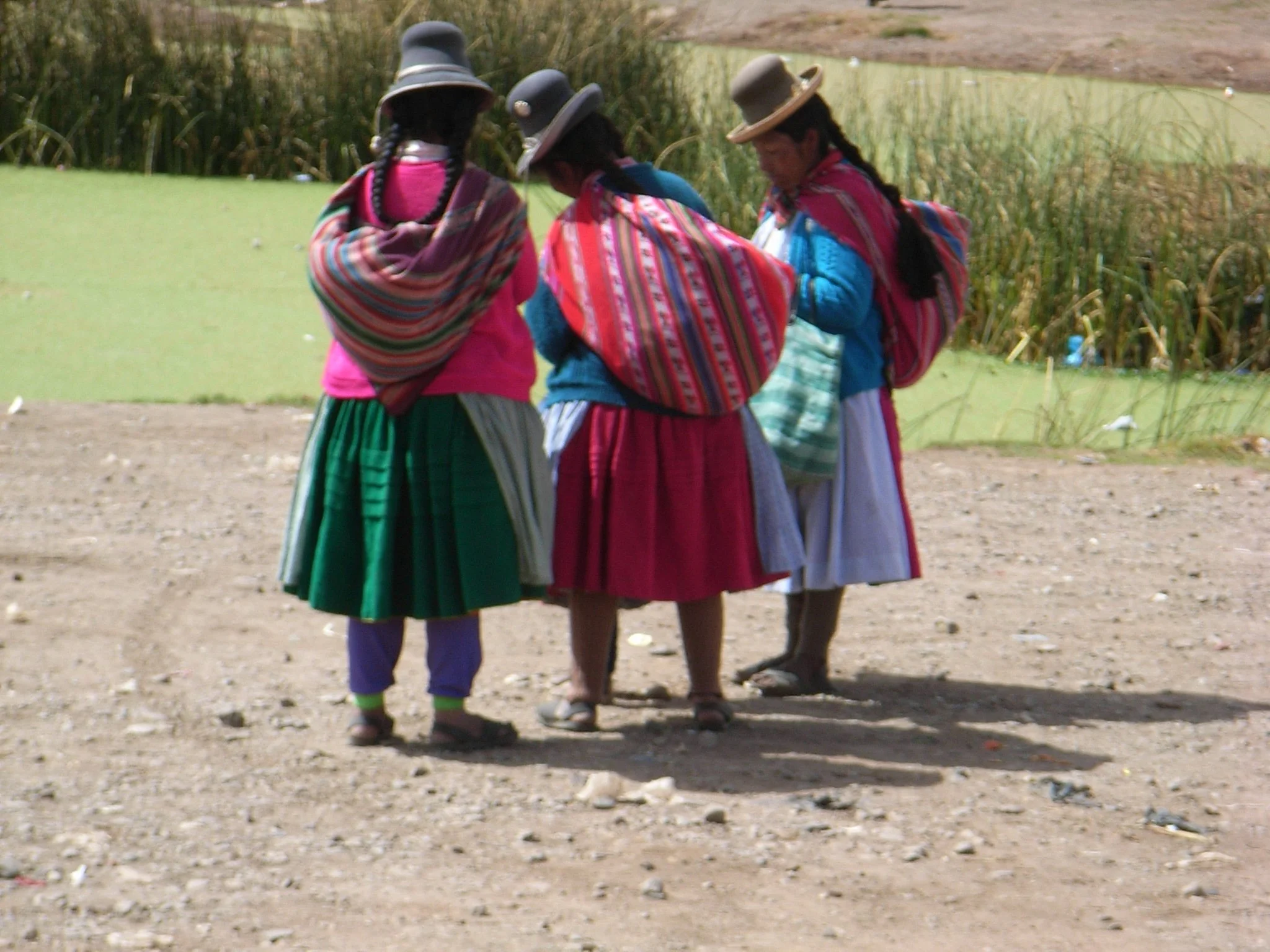 Vijf vrouwen in kleurrijke traditionele kleding en hoeden, staan samen op een landbouwveld, met een meer en rijstvelden op de achtergrond.