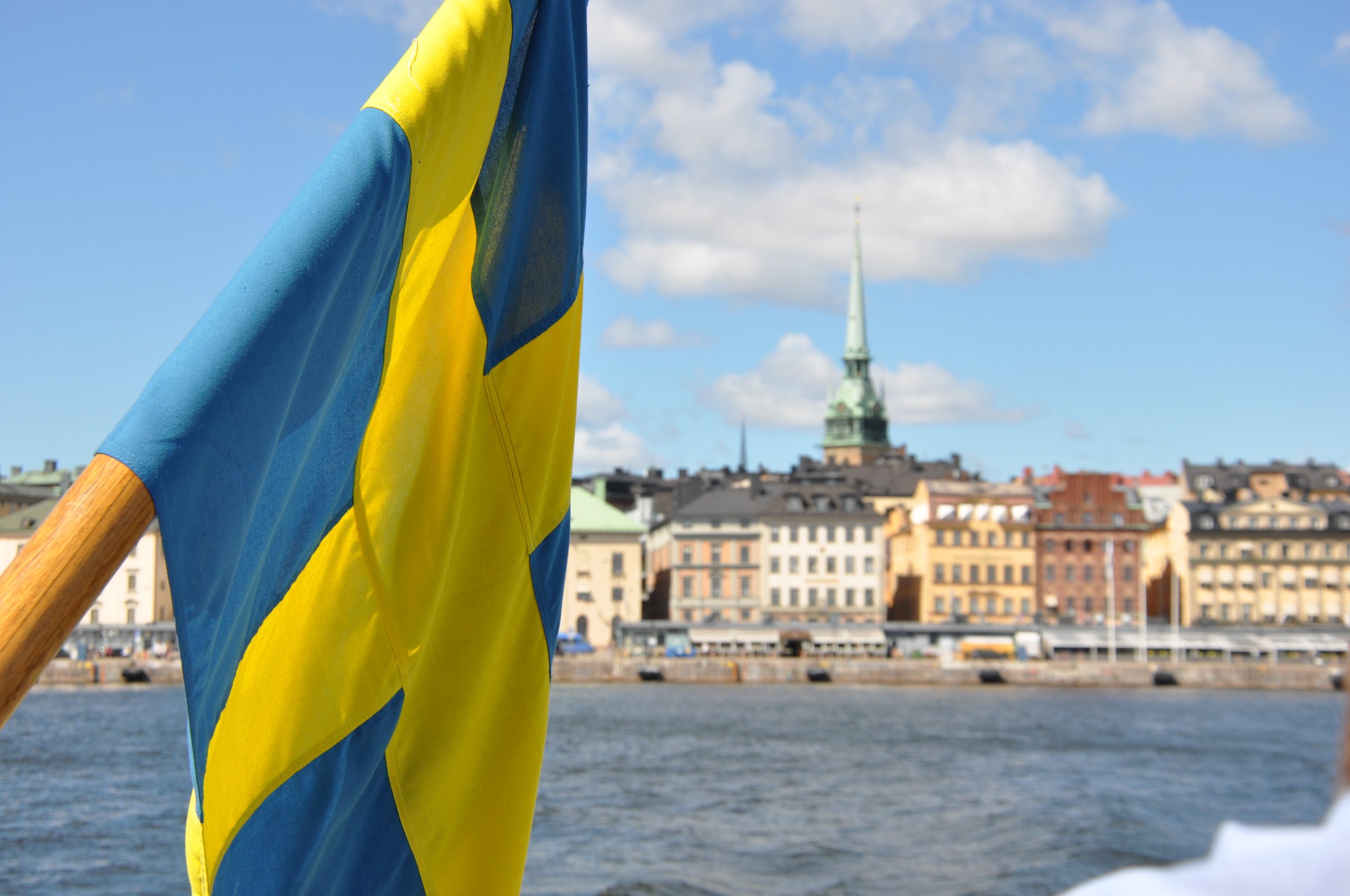 De afbeelding toont een blauwe en gele Zweedse vlag voor een achtergrond van een stadsgezicht met water, oude gebouwen en een kerktoren onder een blauwe hemel met wat witte wolken.