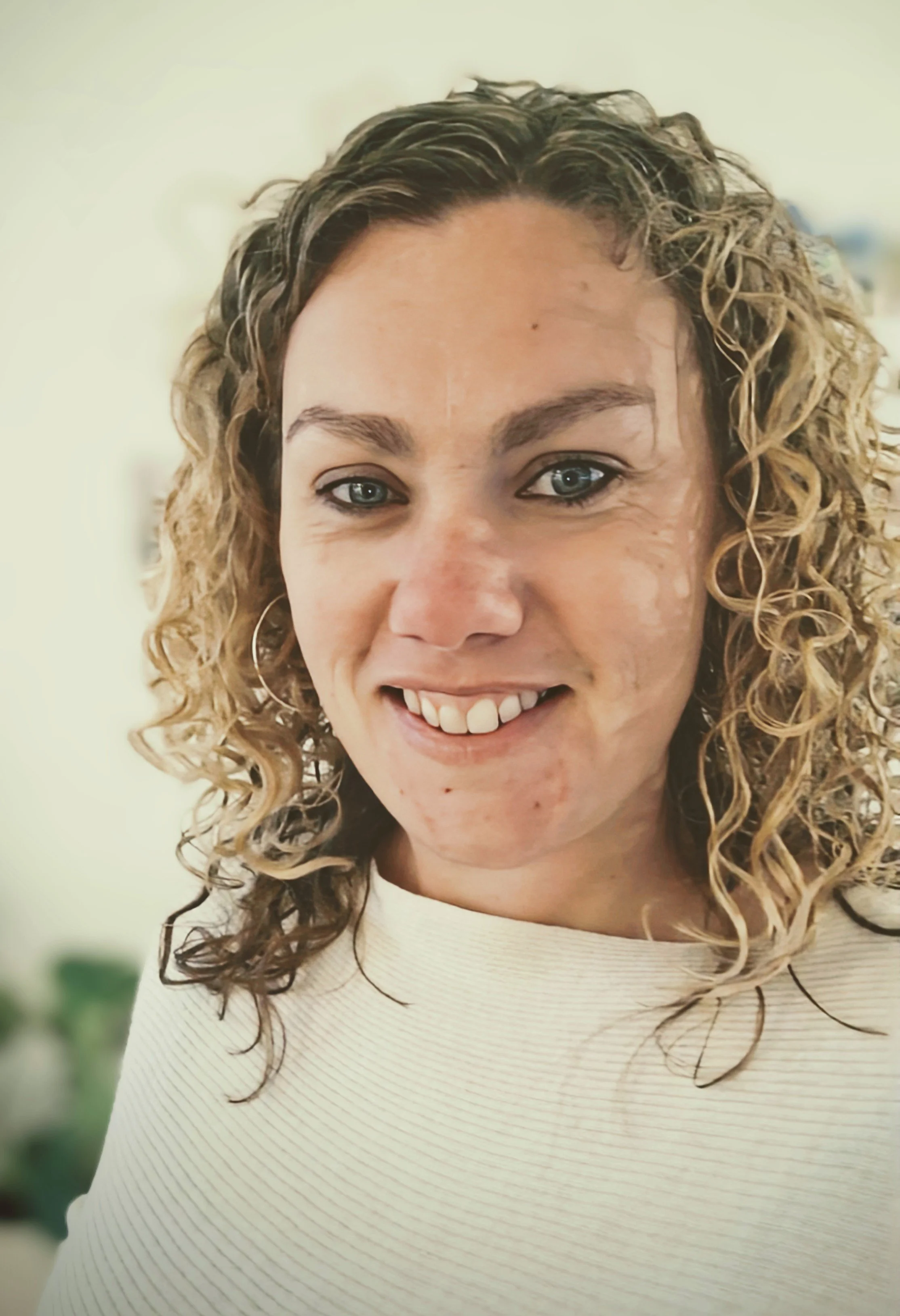 Close-up van een lachende vrouw met blauwe ogen en gekrulde lichtbruine haar, draag een witte trui.