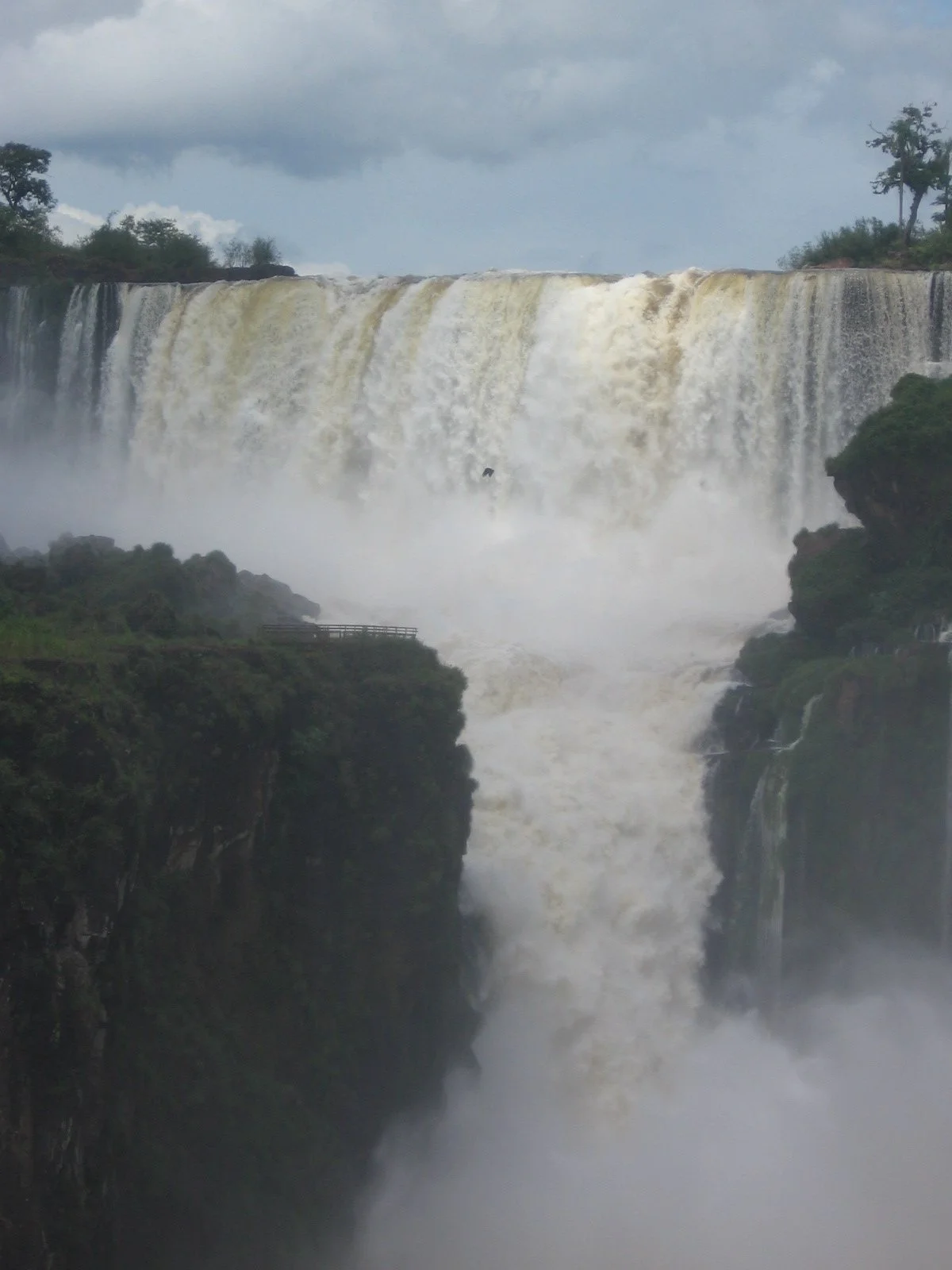 Iguacu waterval met grote hoeveelheid water die neerkomt, omgeven door groene rotsen en bomen, met een bewolkte hemel erboven.