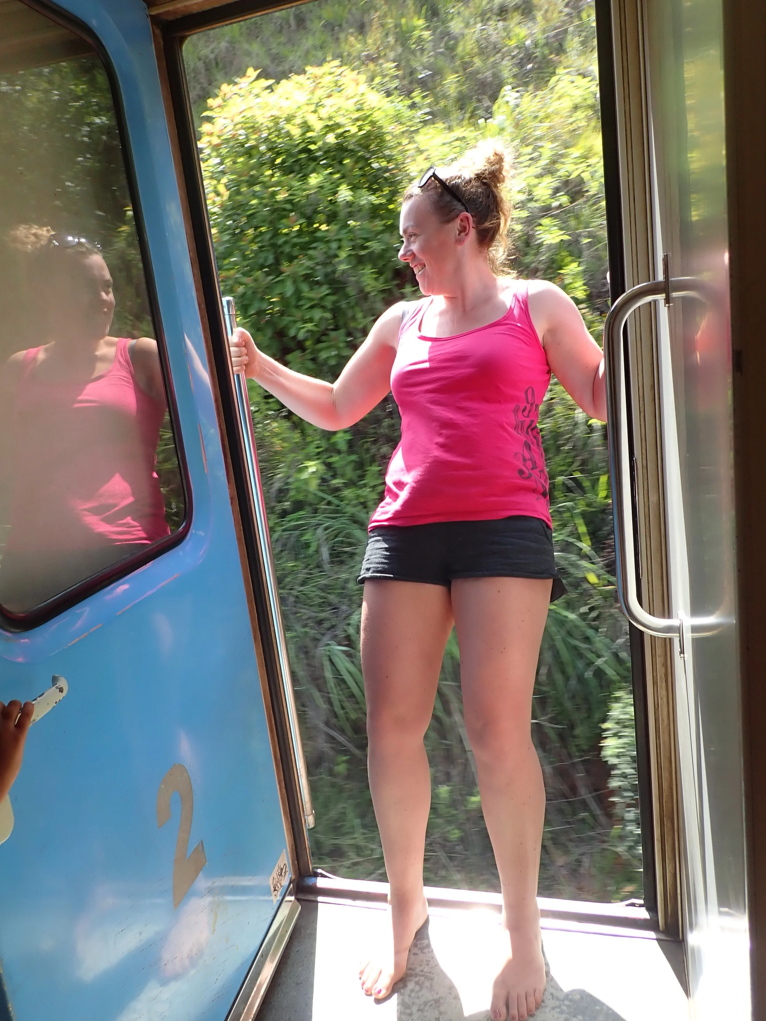 Vrouw in een roze top en zwarte shorts staat op een trambetransfer, lachend, terwijl ze de deur vasthoudt, met een groene natuurlijke achtergrond buiten.