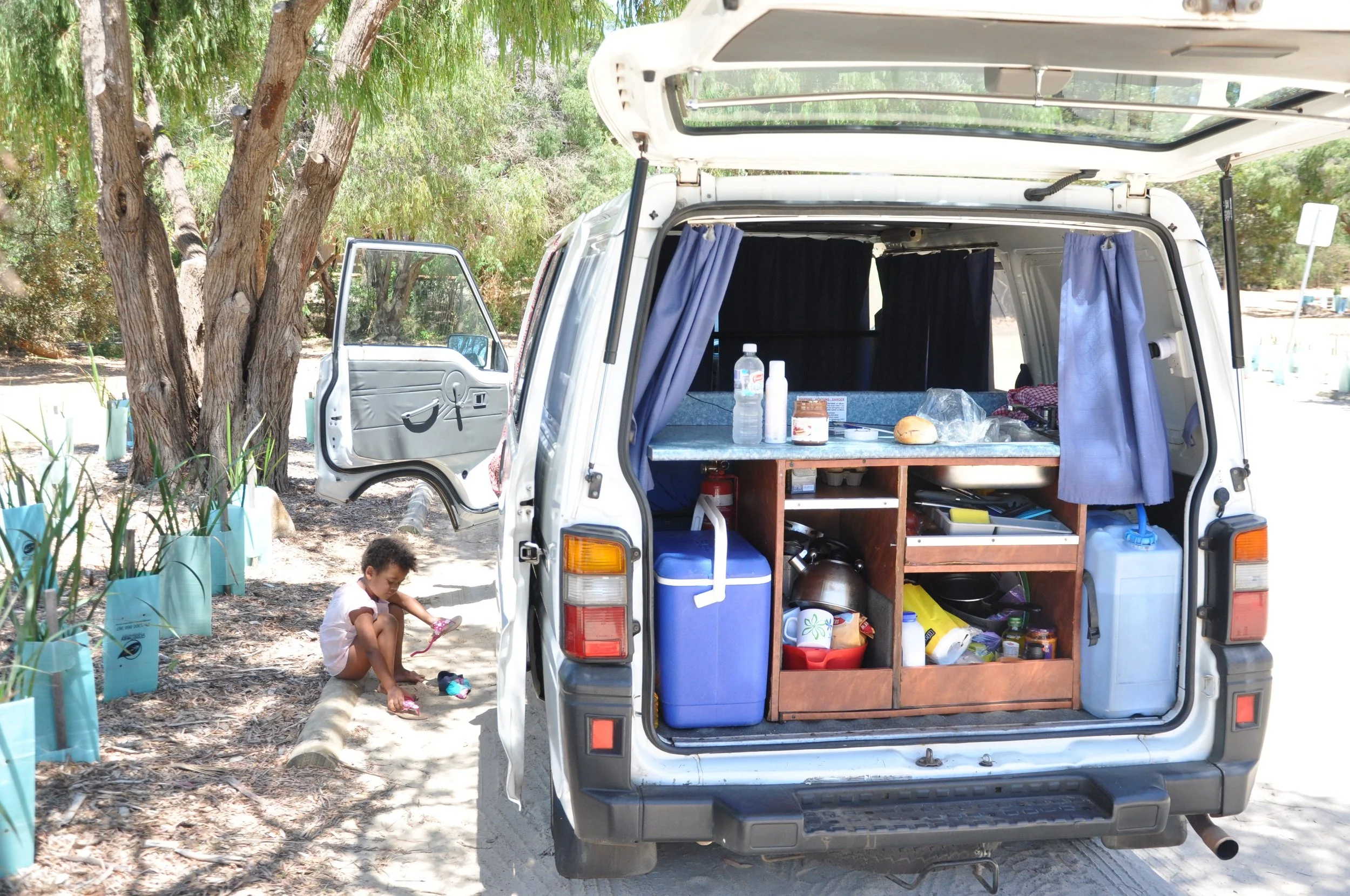 Een camper met open achterdeuren die is ingericht voor kamperen, met een kind zittend op de grond naast de camper, in een natuurlijke omgeving met bomen.