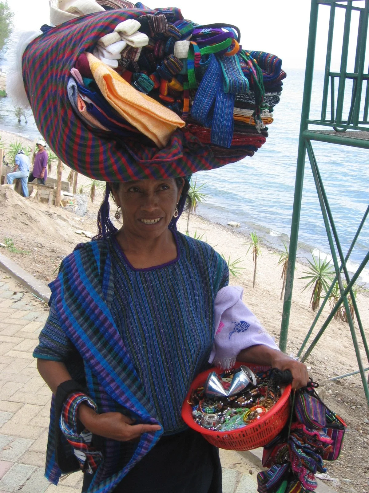 Een vrouw die traditionele kleding draagt en een grote kop met kleurrijke textiles. Ze houdt een rood mandje met sieraden en accessoires in haar hand en staat naast een strand.