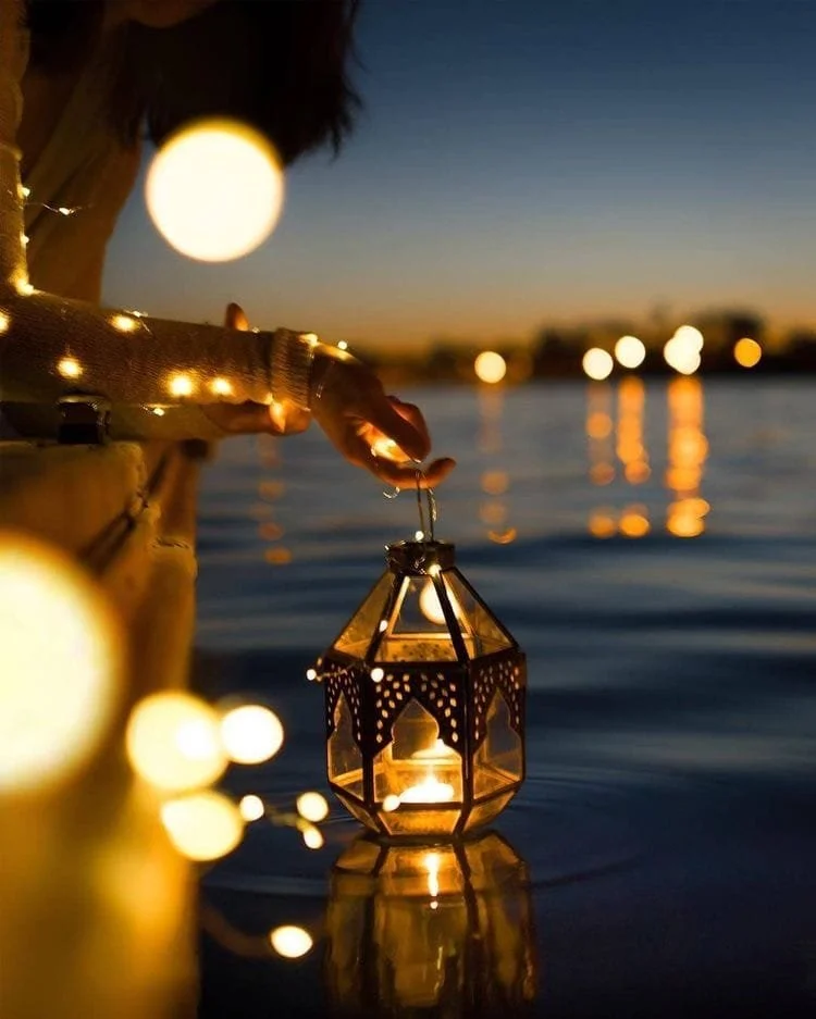 Une personne tient une lanterne en verre suspendue au-dessus d'un corps d'eau lors du crépuscule, avec des lumières suspendues qui se reflètent dans l'eau.