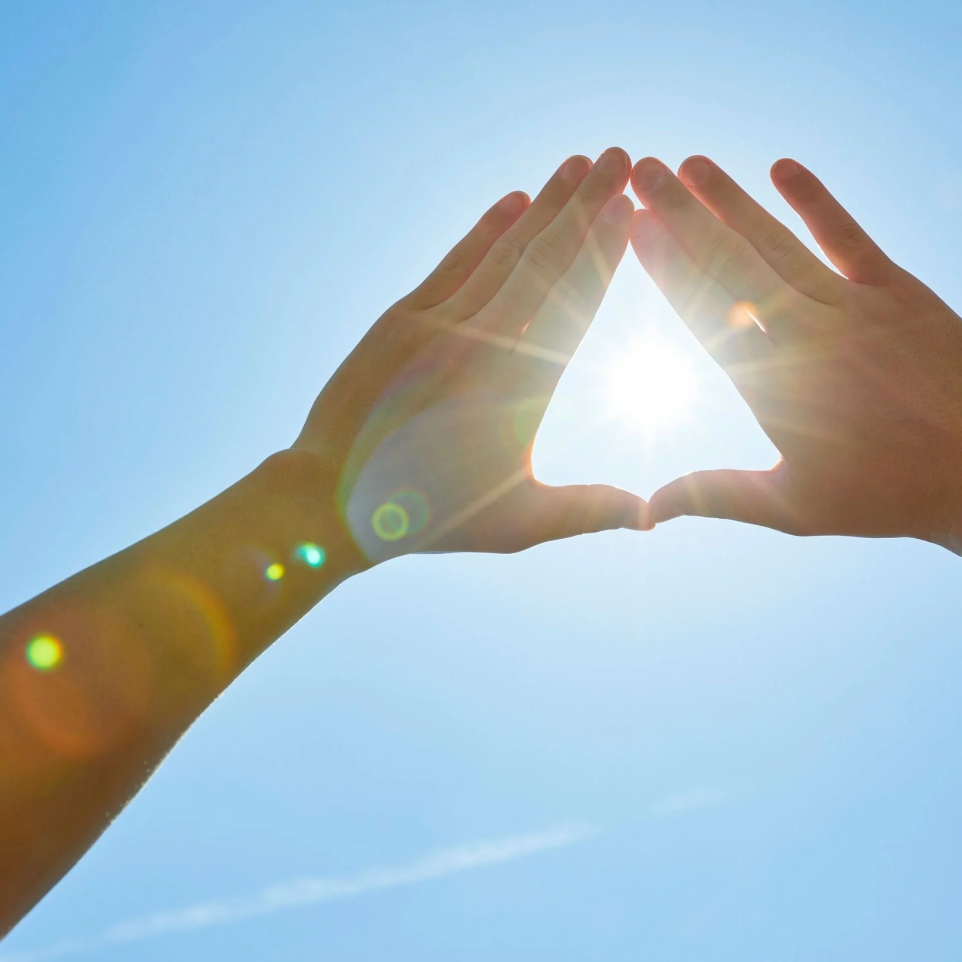 Mains formant un cœur avec le soleil brillant derrière, dans un ciel bleu clair.