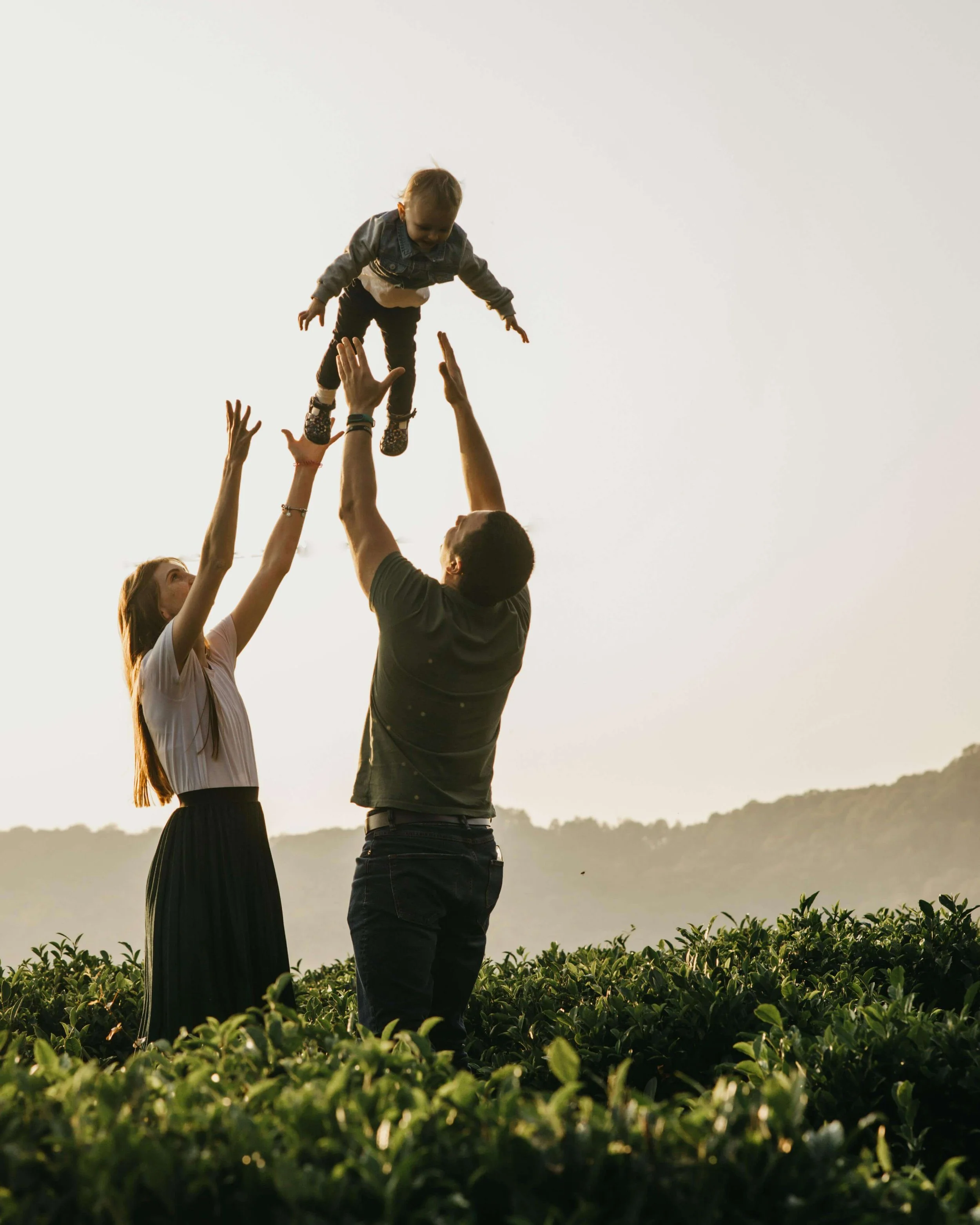 Un groupe de trois personnes dans un champ vert, levant un enfant en l'air lors d'un coucher de soleil.