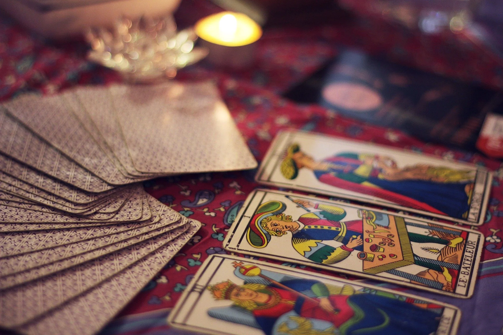 Jeu de tarot avec plusieurs cartes disposées sur une surface en tissu rouge à motifs colorés, un paquet de cartes face cachée à gauche, et une petite bougie allumée en arrière-plan.