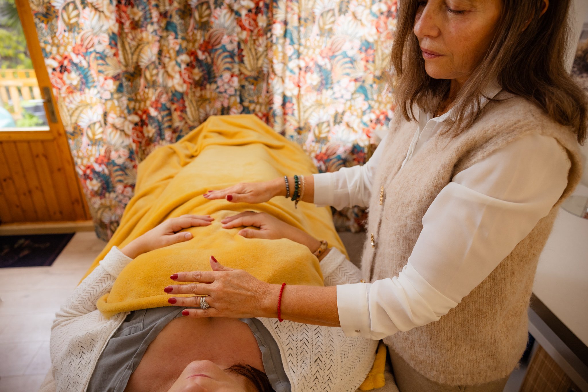 Une femme donne un massage à une jeune fille allongée sur une table, dans une pièce décorée avec des rideaux fleuris et un intérieur chaleureux.