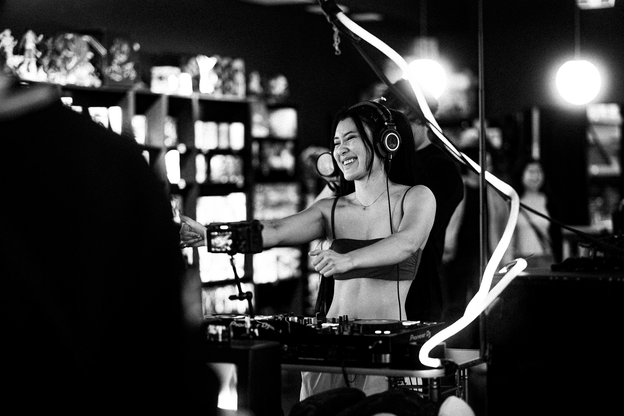 A woman smiling and dancing behind DJ equipment, wearing headphones, in a lively indoor setting with shelves and lighting.