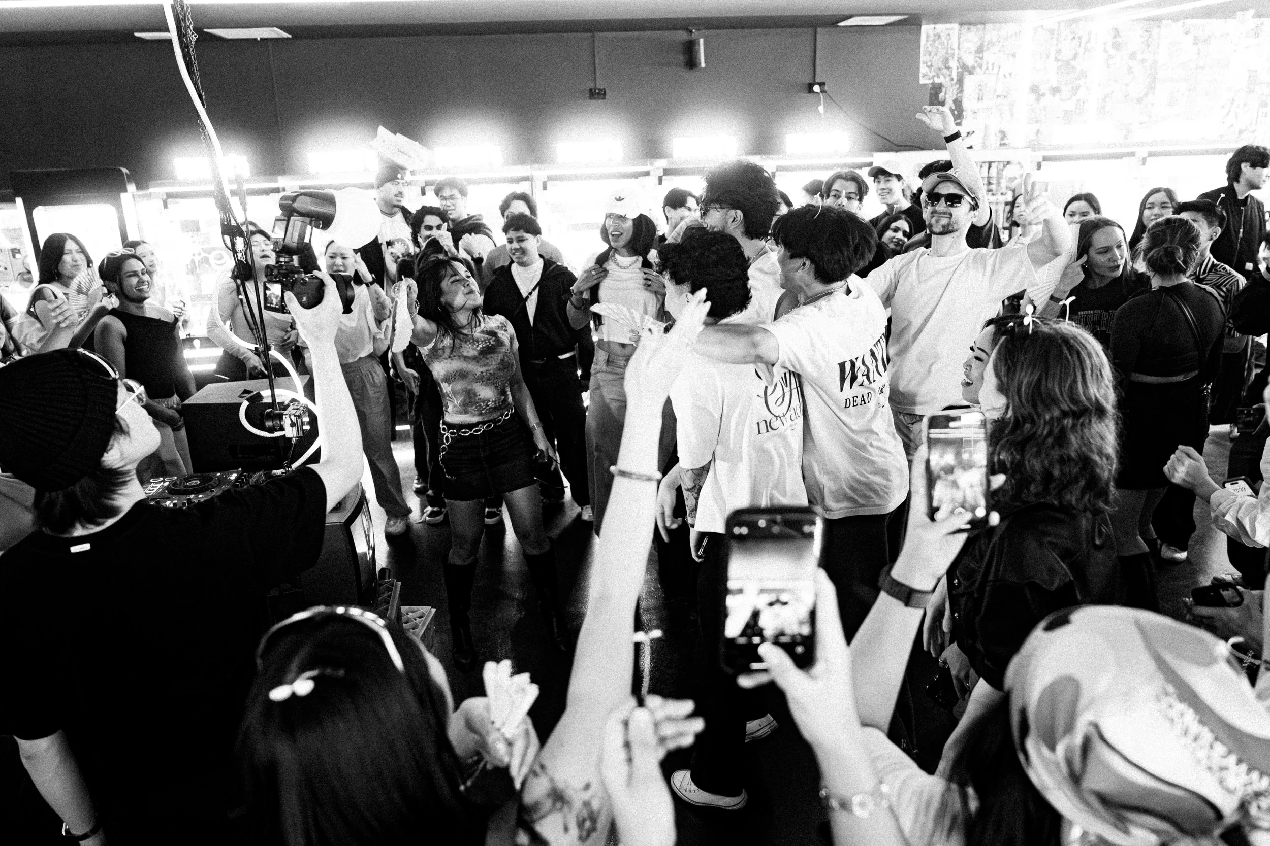 A group of people dancing and taking photos at a lively indoor event, with some individuals in the center dancing and others capturing the moment on their phones.
