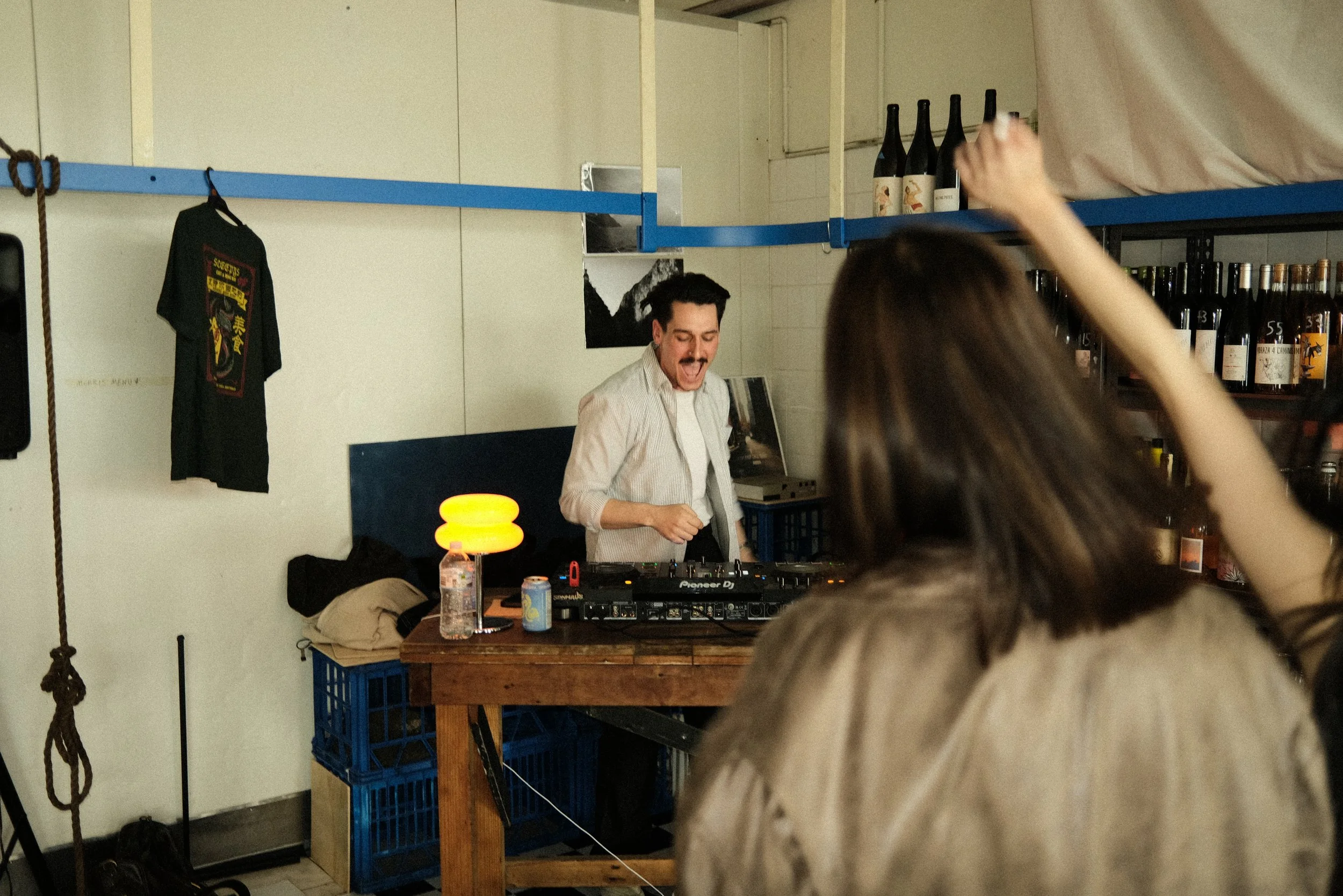 A DJ with dark hair and a mustache is playing music at a party, standing behind a DJ console on a wooden table. A woman with brown hair is dancing with her arm raised in front of him. The background includes shelves with bottles and a black t-shirt hanging on the wall.
