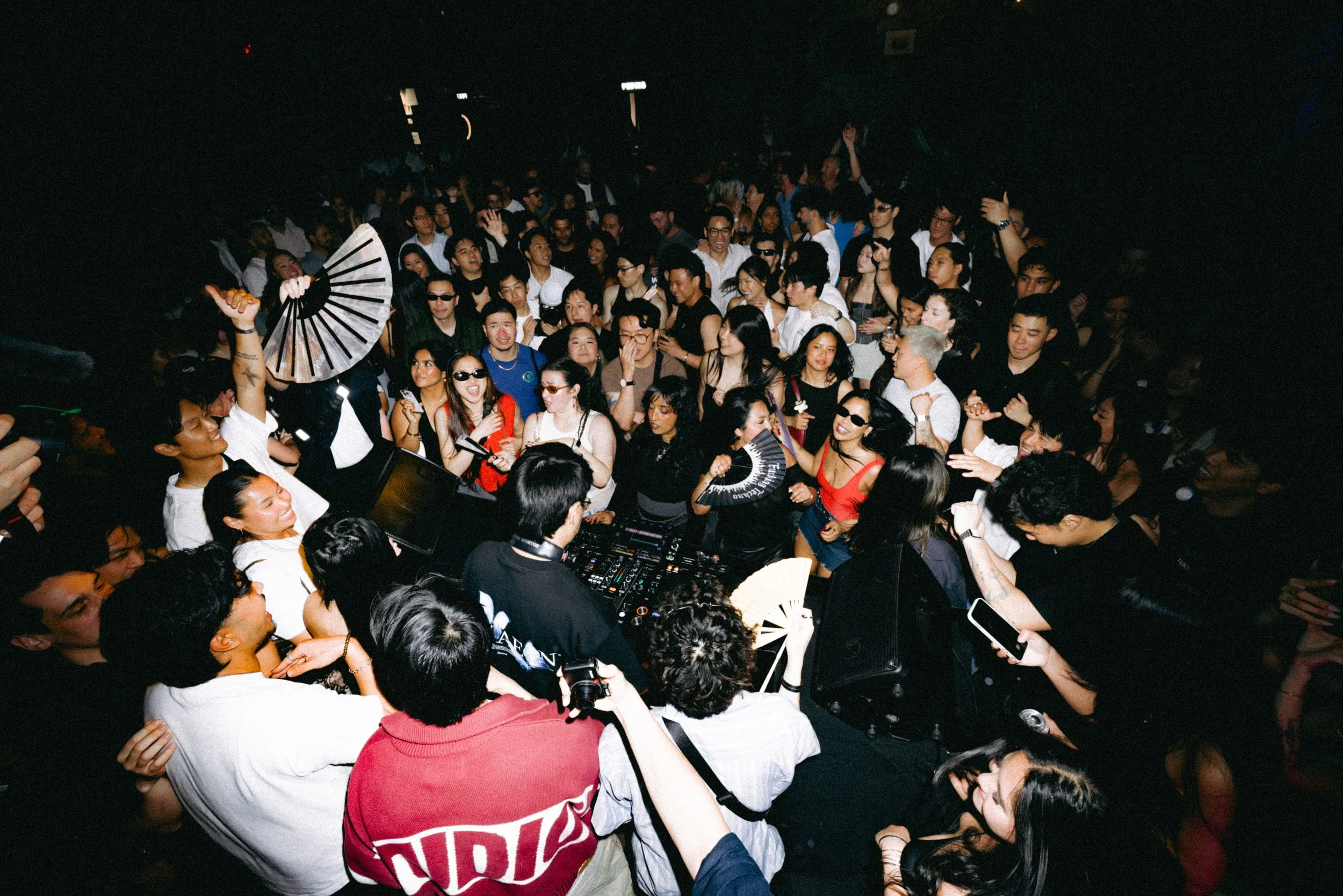 Crowd of people dancing at a nightclub with a DJ playing music in the center.