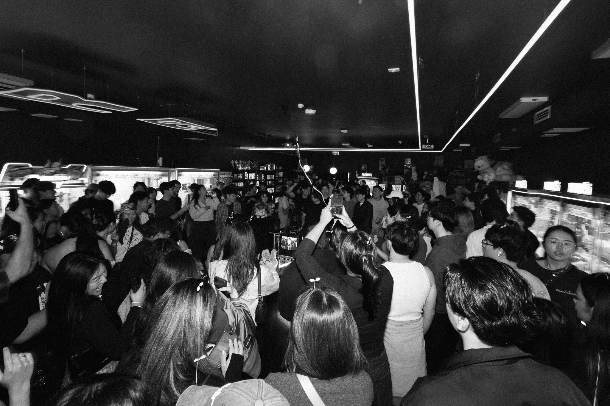 A crowd of people gathered in a room with shelves of toys and vending machines. Some are taking photos, and there are stuffed animals on top of the shelves. The room has a dark ceiling with bright light strips.