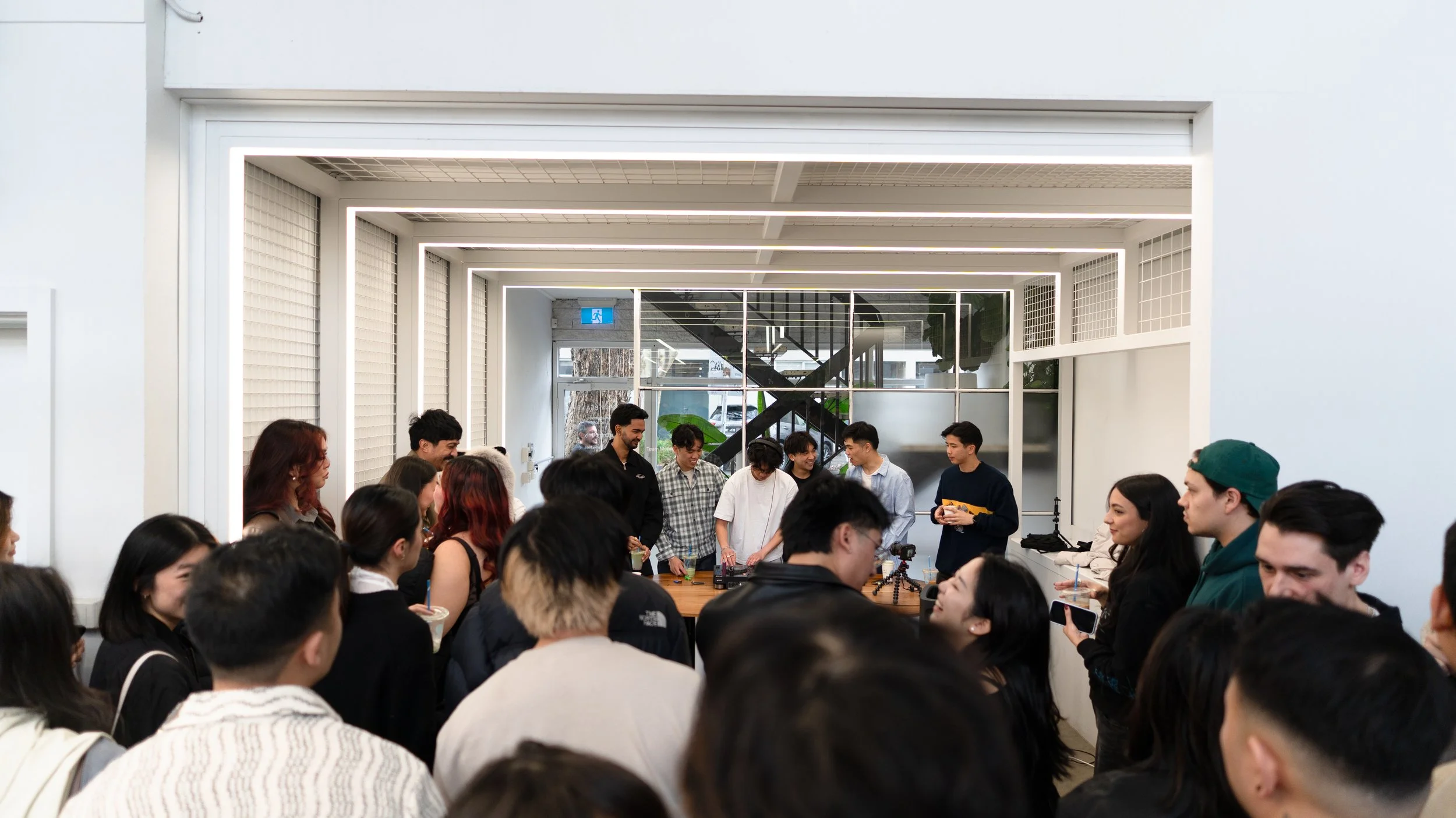 Group of people gathered in a modern interior space with large glass windows and a staircase in background, participating in a workshop or presentation.