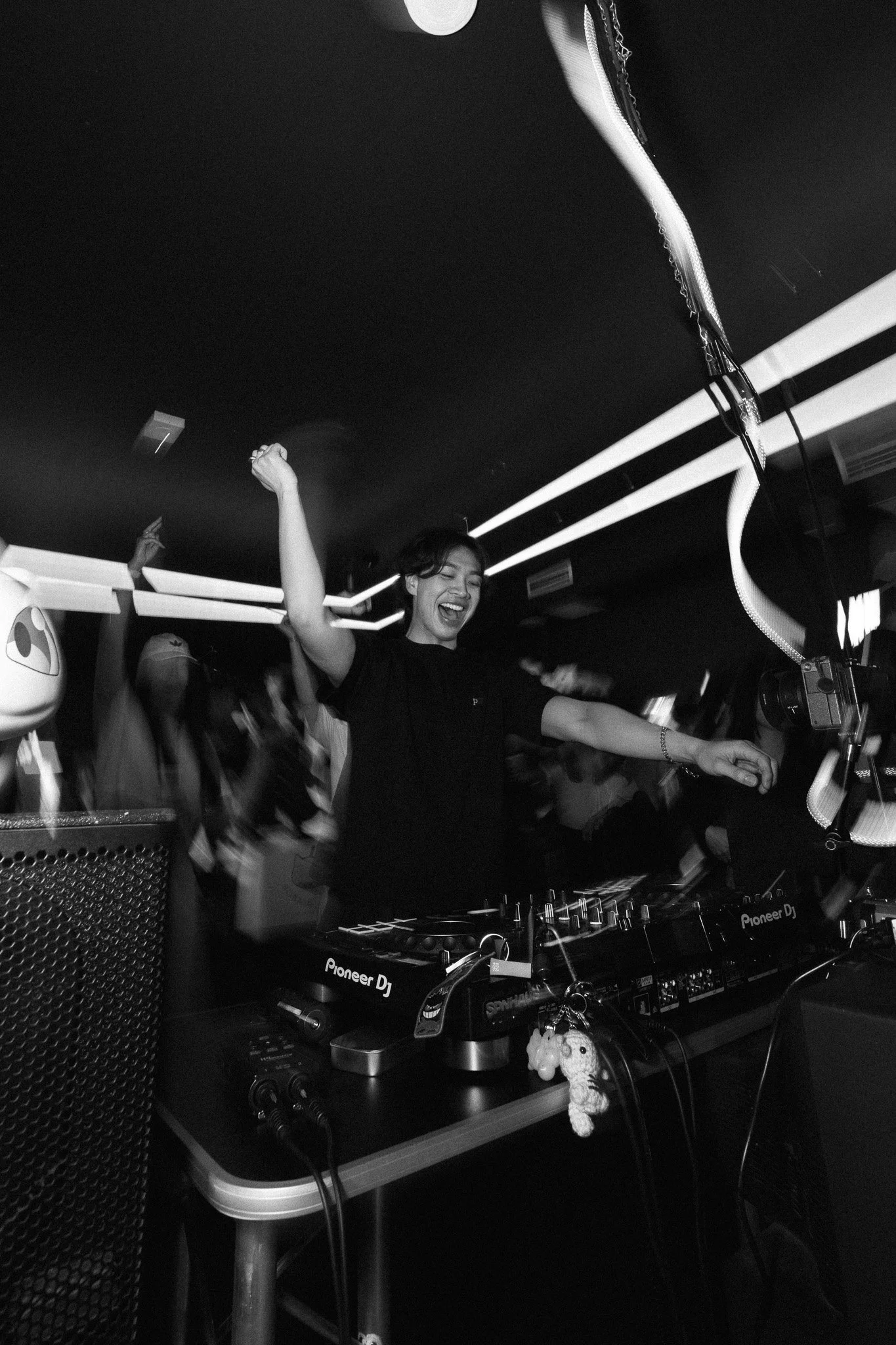 A woman DJ smiling and dancing at a club with a DJ setup, surrounded by party-goers, in black and white.