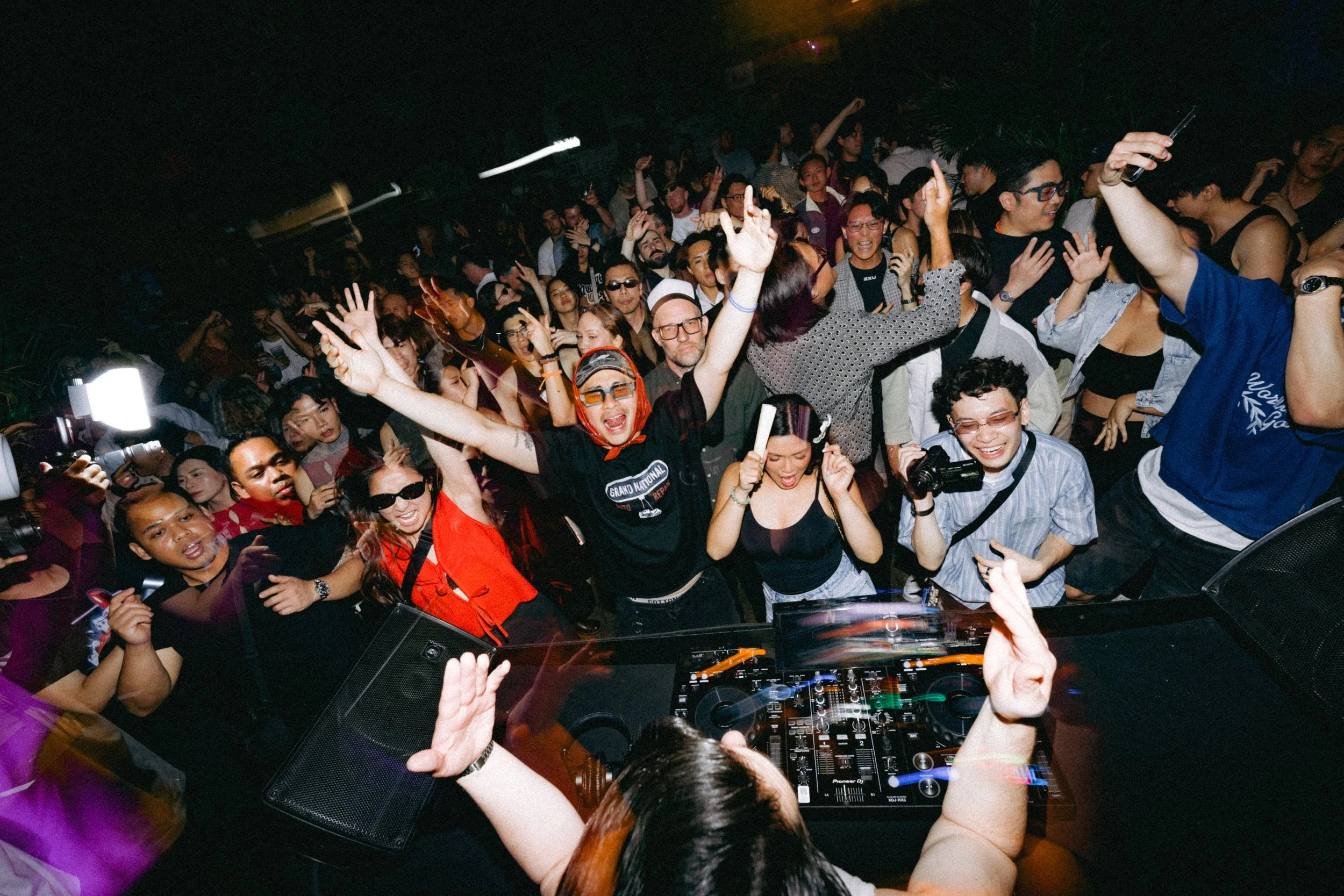 A crowded nightclub dance floor with people dancing and enjoying the music, viewed from behind a DJ at a DJ booth.
