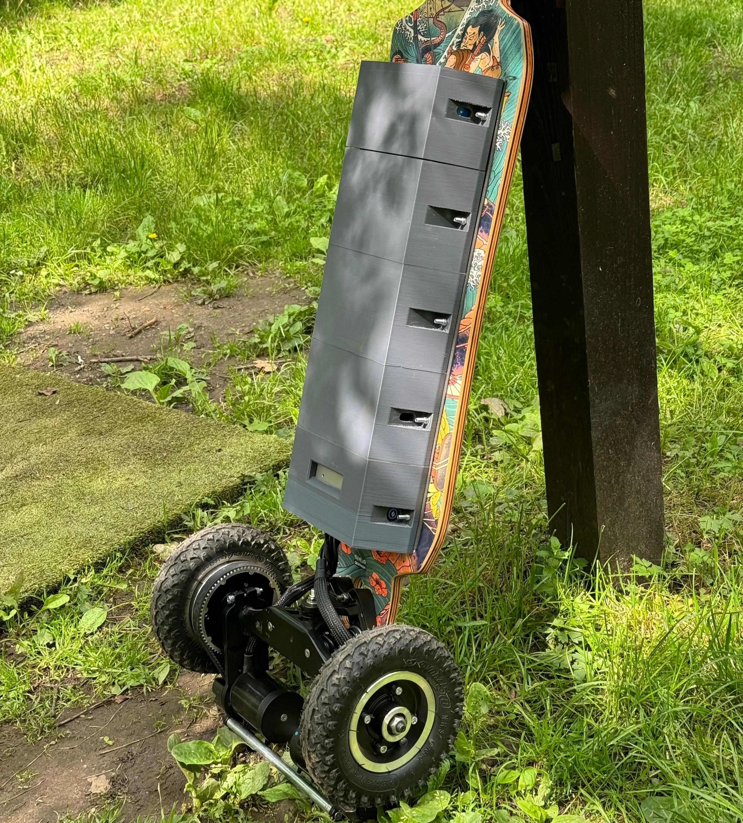 An electric skateboard with large wheels leaning against a black wooden post in a grassy outdoor area.