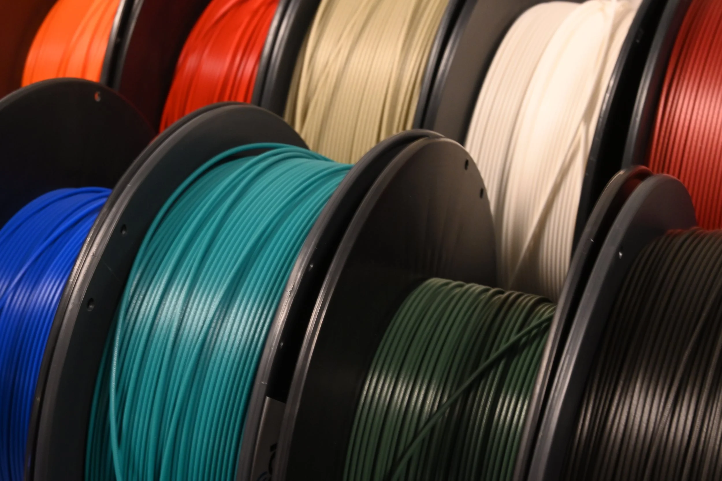 Spools of various colored 3D printer filament in black, orange, beige, red, blue, teal, and green.