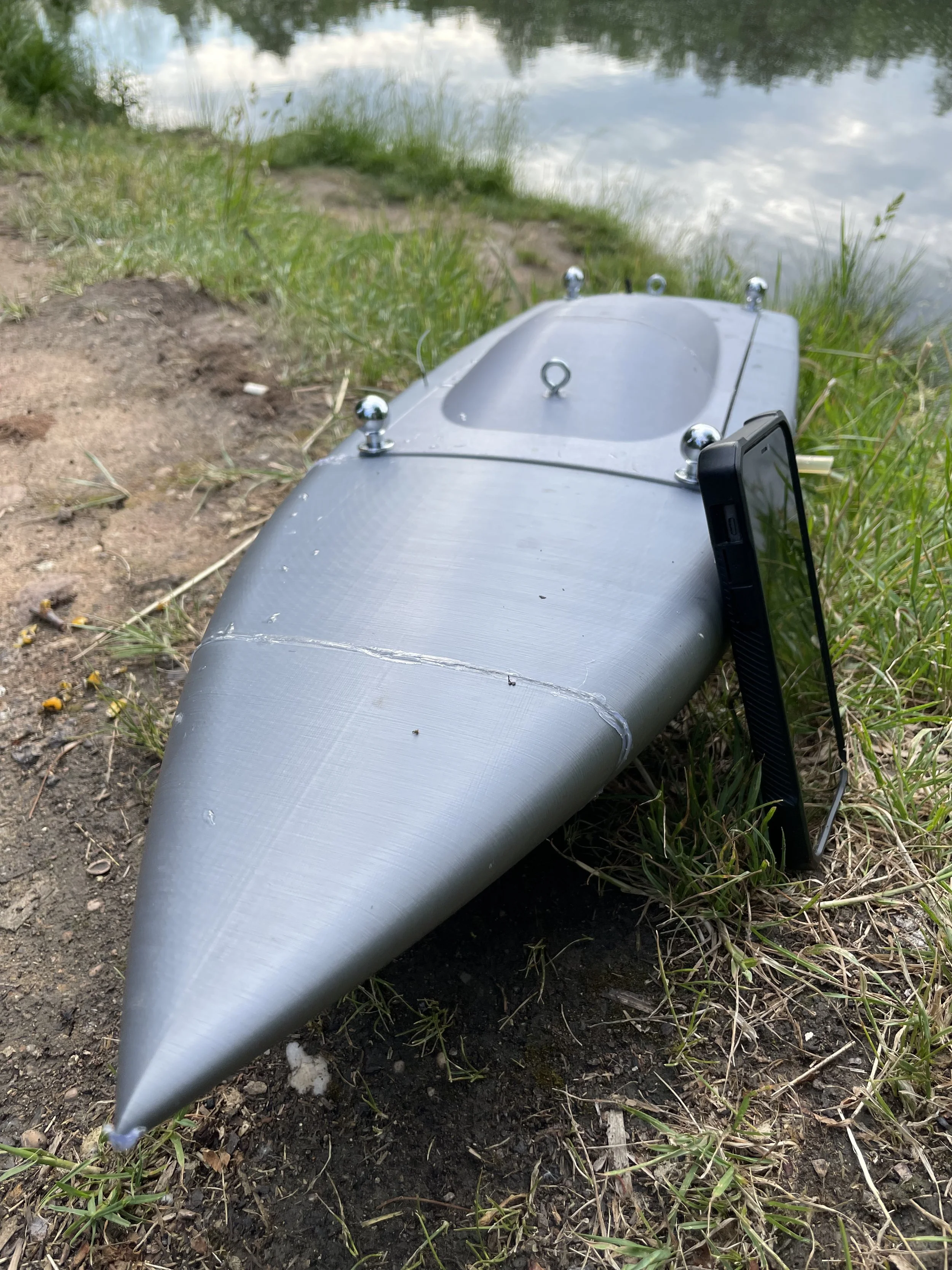 A small black robot boat on the ground near a body of water, with a smartphone propped against it.