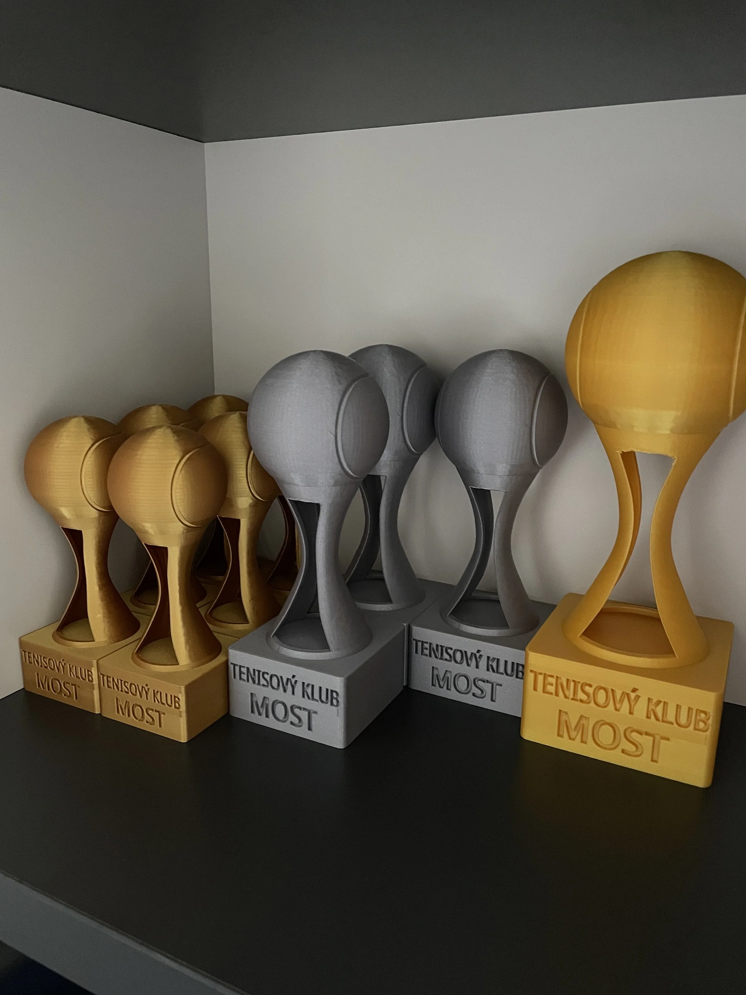 Three sets of brightly colored, modern-looking trophies or awards in yellow, gray, and yellow, placed on a black shelf against a white wall, with inscriptions in a foreign language.