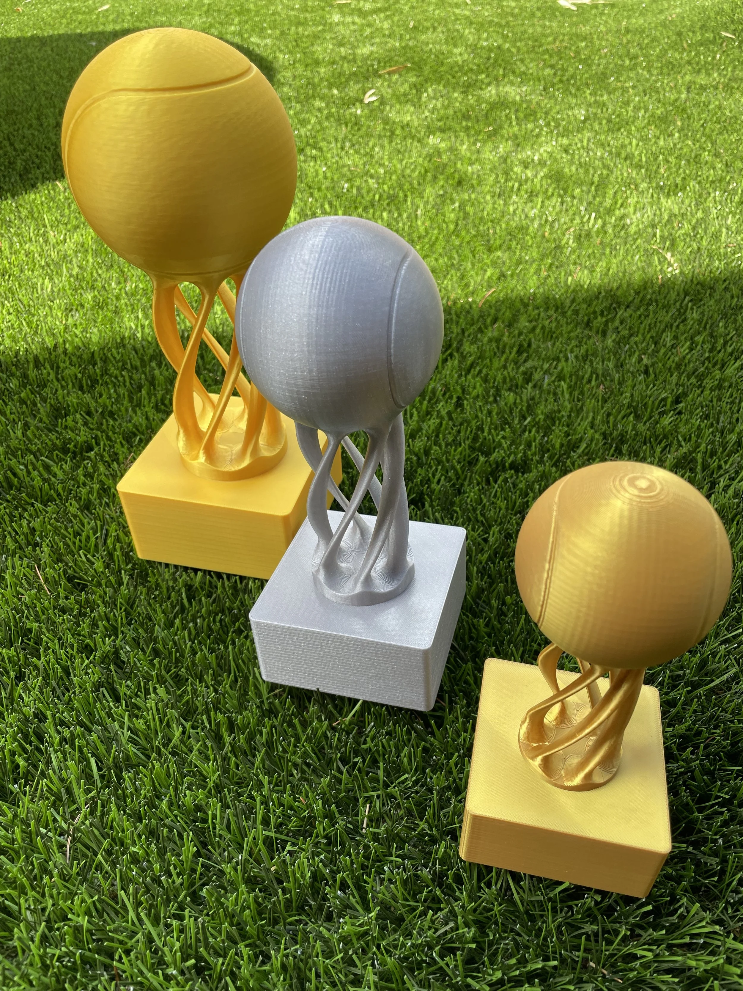Three trophy-style awards on a grassy surface, with two gold-colored trophies and one silver-colored trophy, each with a globe on top connected by loops, mounted on colored bases.