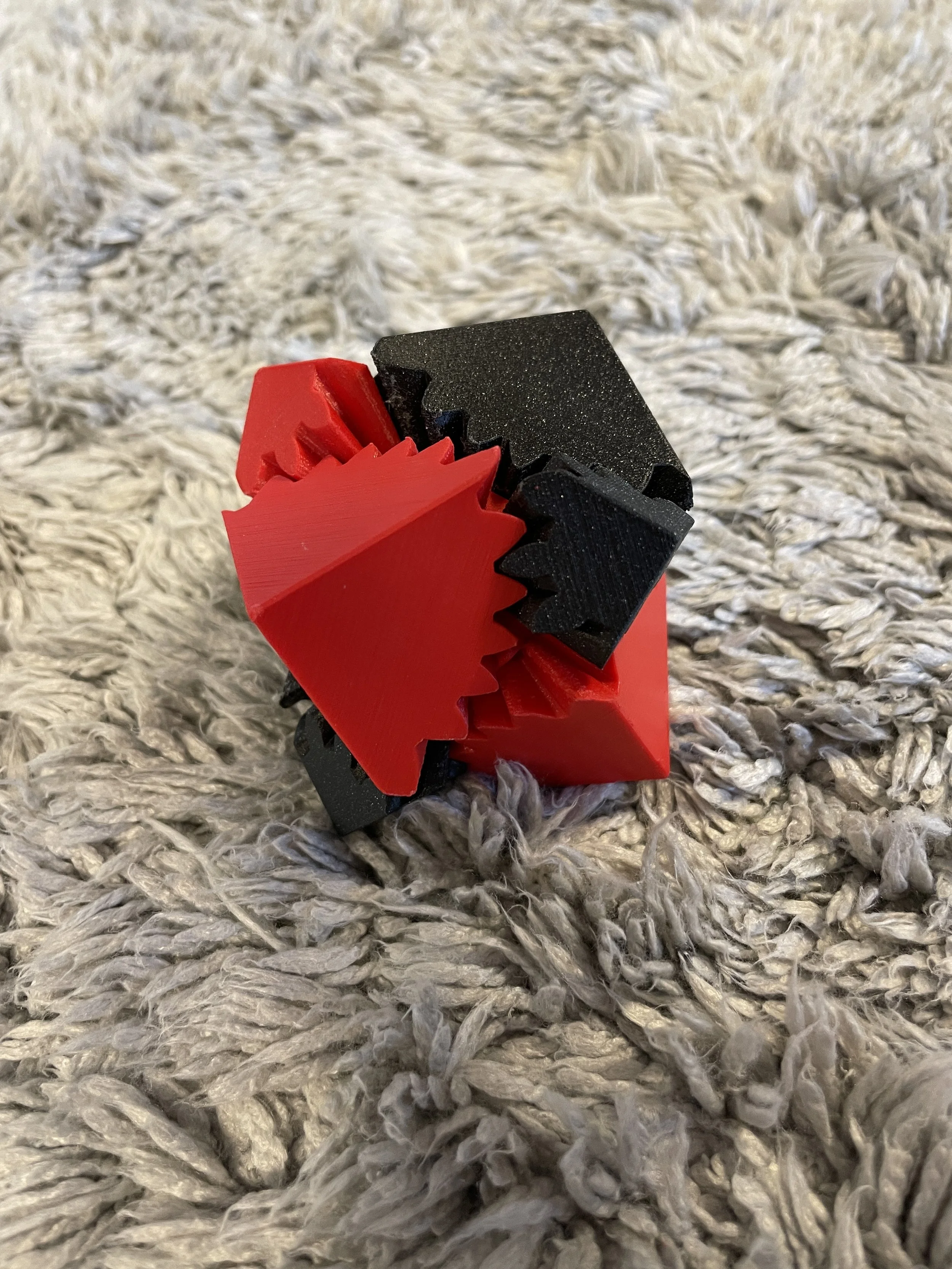 A close-up of a 3D-printed puzzle made of interlocking black and red pieces, resting on a textured, gray faux-fur rug.