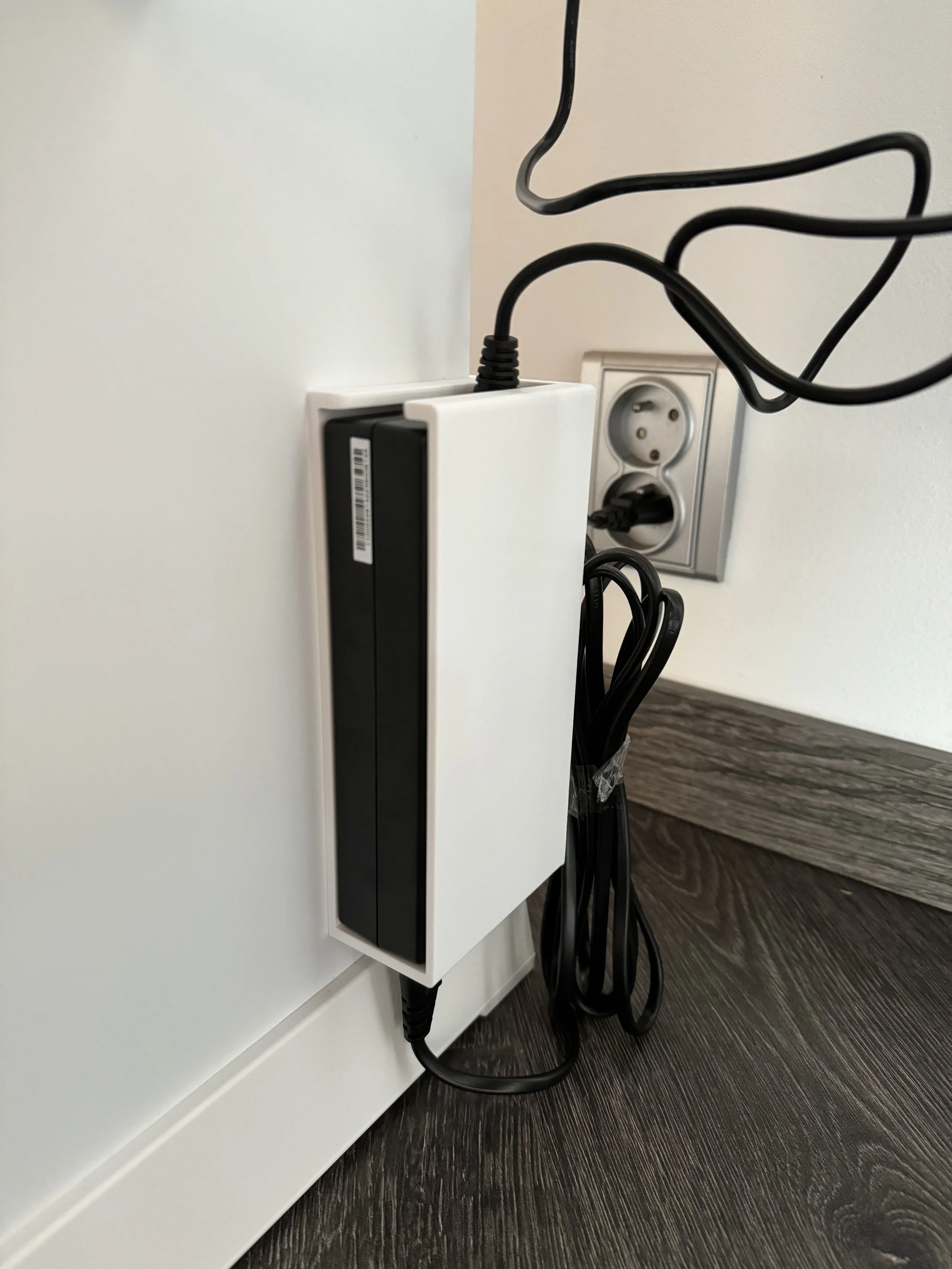 A white Wi-Fi router plugged into a power outlet, with black cables connected and hanging down, on a dark wood floor next to a white wall and baseboard.
