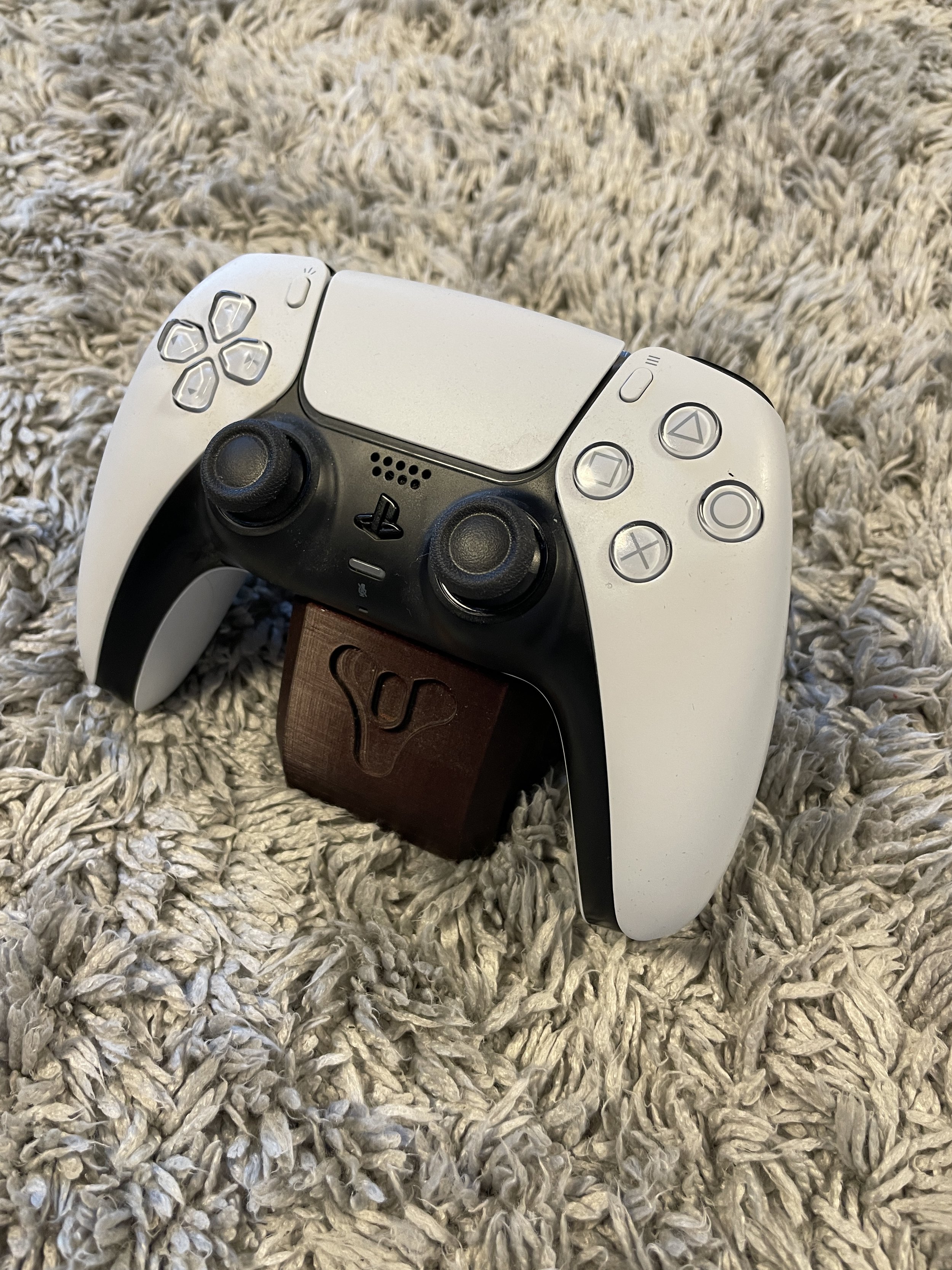 A white PlayStation 5 DualSense controller resting on a gray shaggy carpet, mounted on a small wooden stand with a logo.