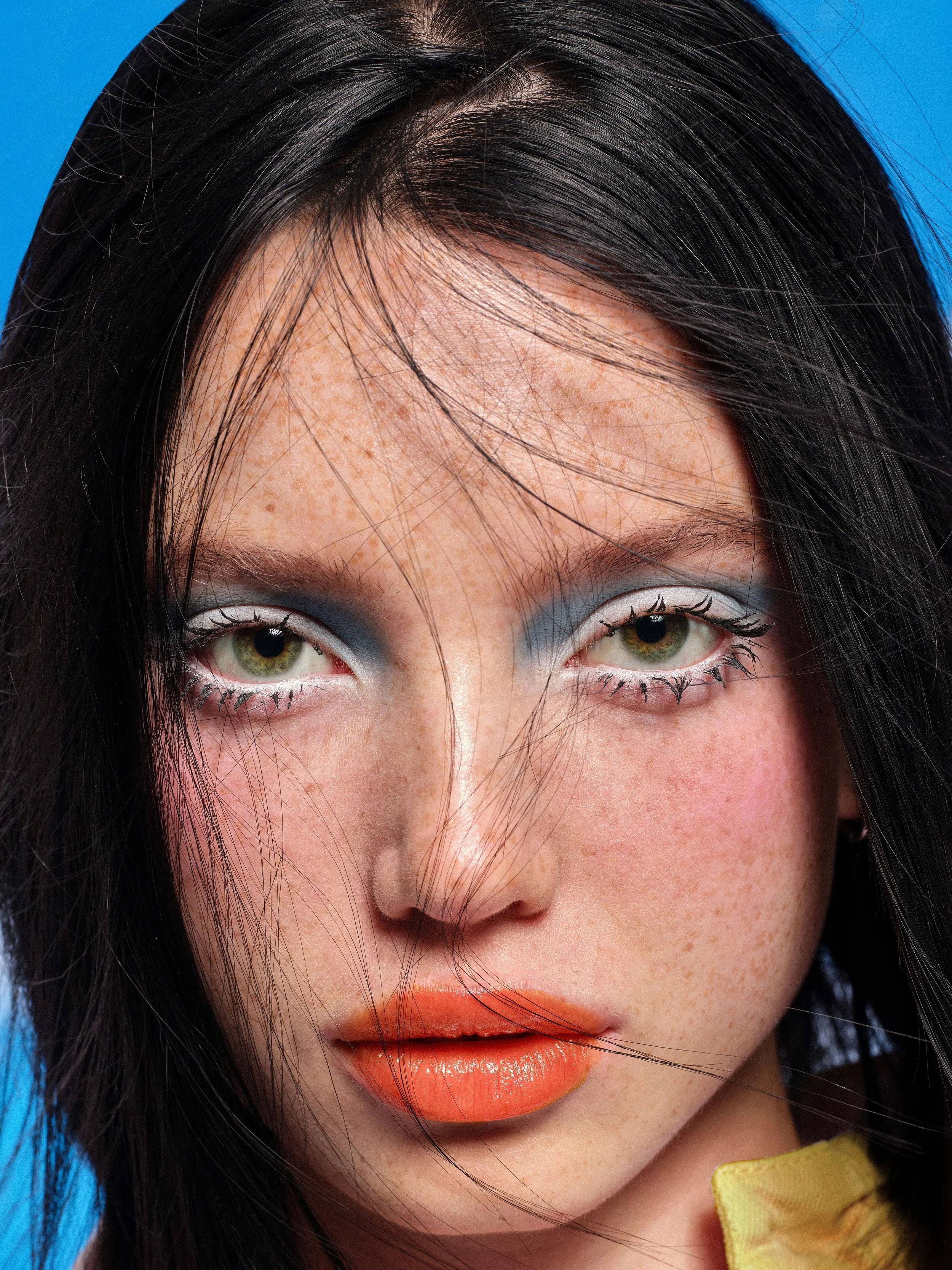 Close-up of a woman with makeup, black hair, and freckles, looking at the camera with a blue background.