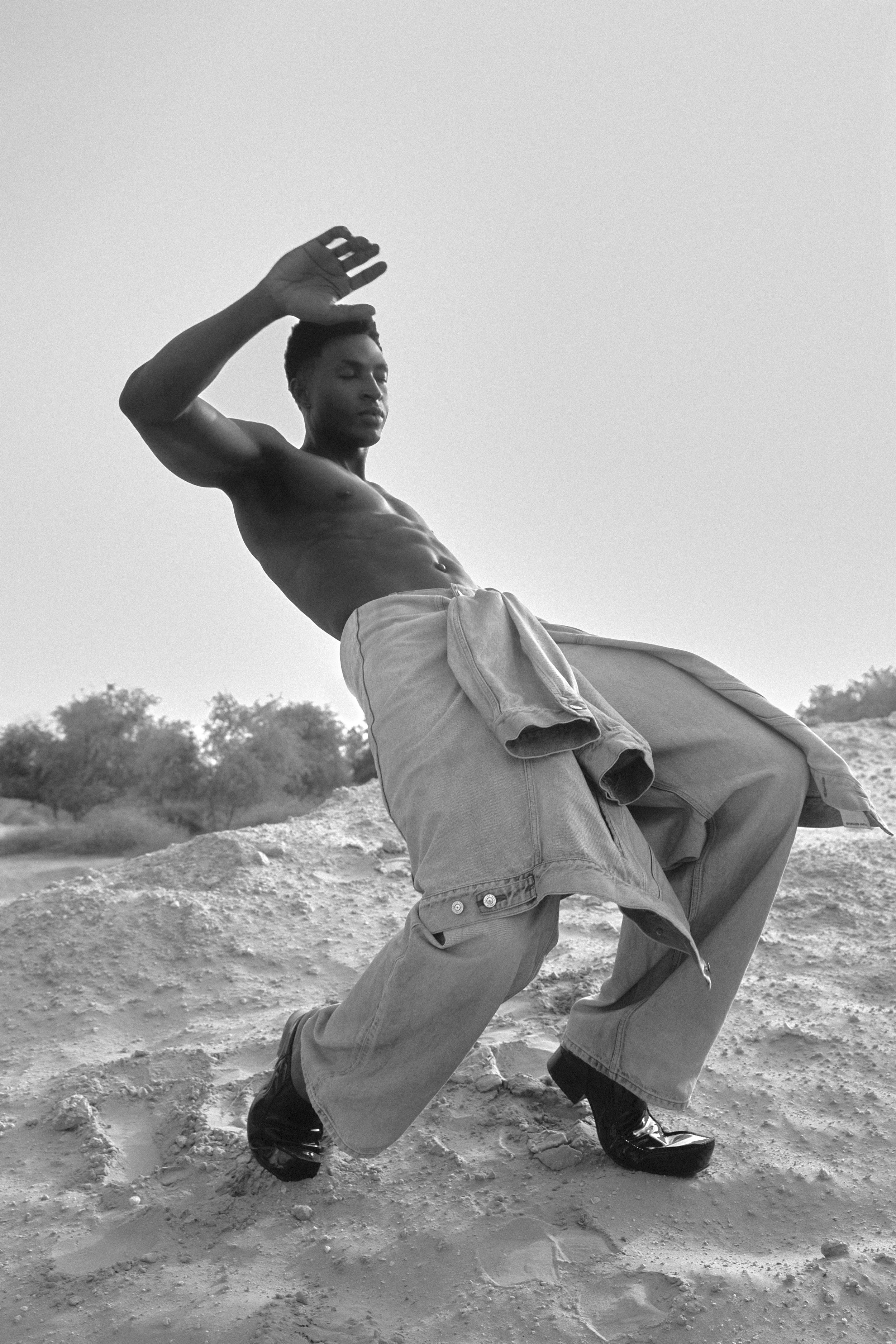 A shirtless young man with an athletic build, wearing baggy pants and shiny black shoes, strikes a pose outdoors on sandy terrain with a clear sky and trees in the background.