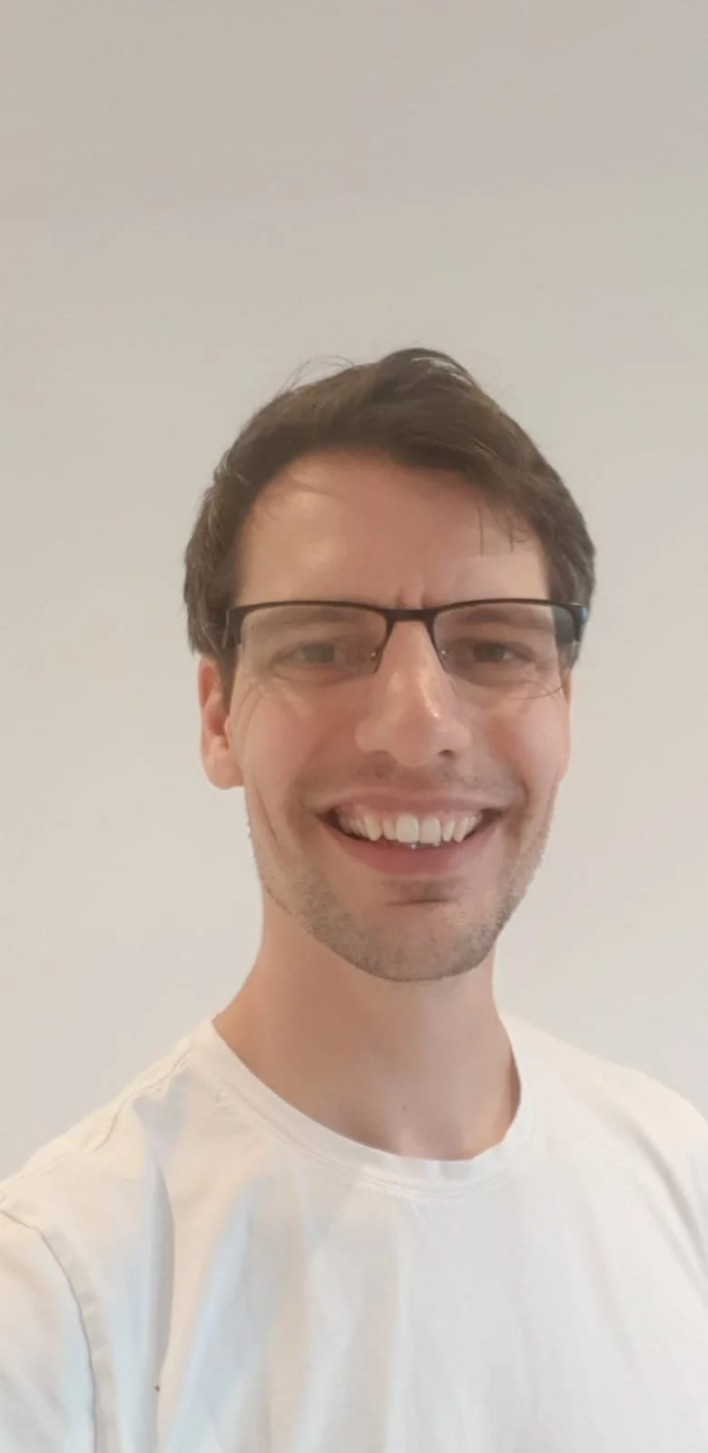 A smiling man wearing glasses and a white t shirt against a plain white background.
