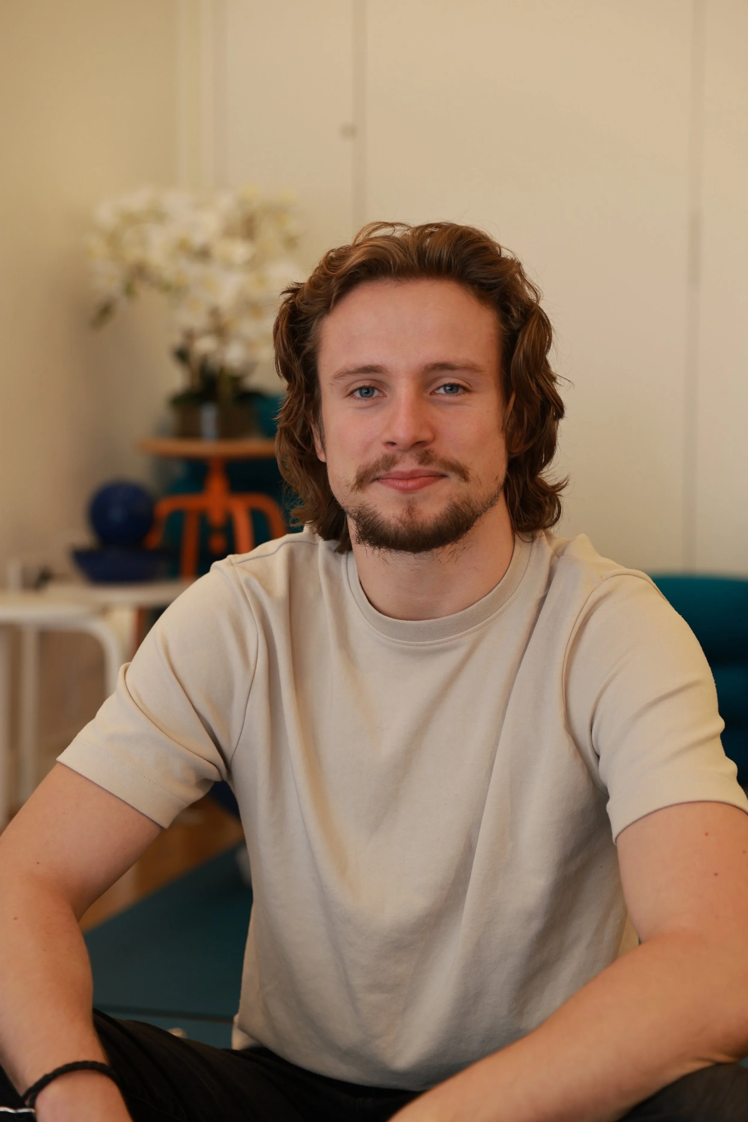 A young man with shoulder-length brown hair, light skin, and a slight beard, sitting indoors with a neutral expression, wearing a beige T-shirt, with plants and furniture in the background.