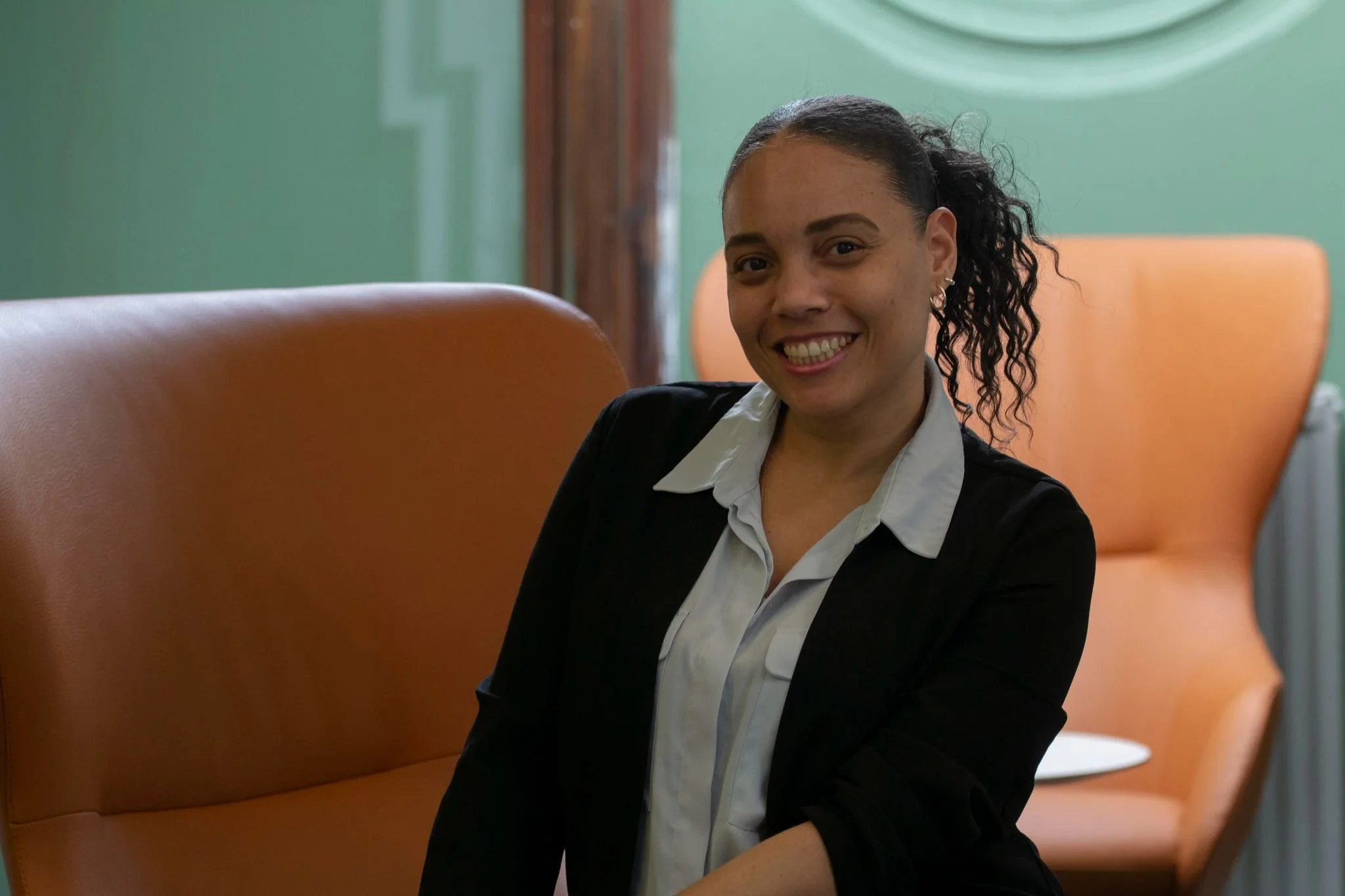 A woman with curly hair tied back, smiling and wearing a black blazer and white shirt, sitting in a modern office or waiting area with green walls and orange chairs.