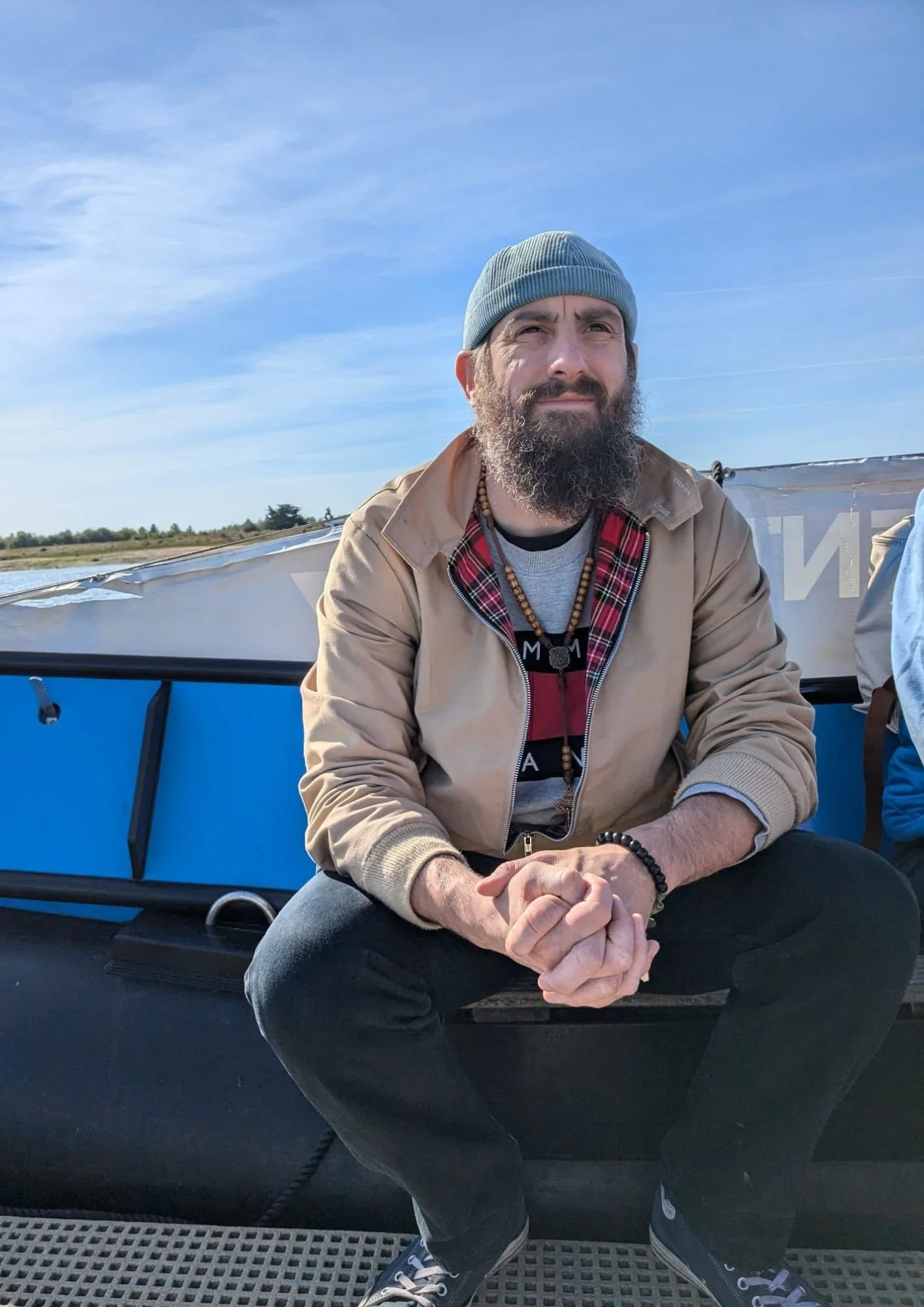 A bearded man wearing a gray beanie, beige jacket, and black pants sitting on a boat with a river and blue sky in the background.