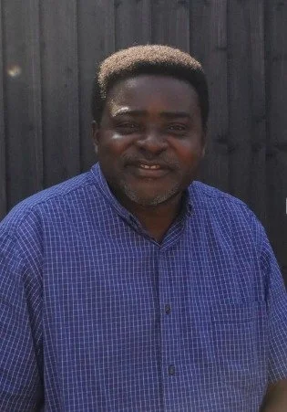 A man with dark skin and a short, light-colored afro hairstyle, wearing a blue checkered shirt, standing outdoors in front of a black wooden fence.