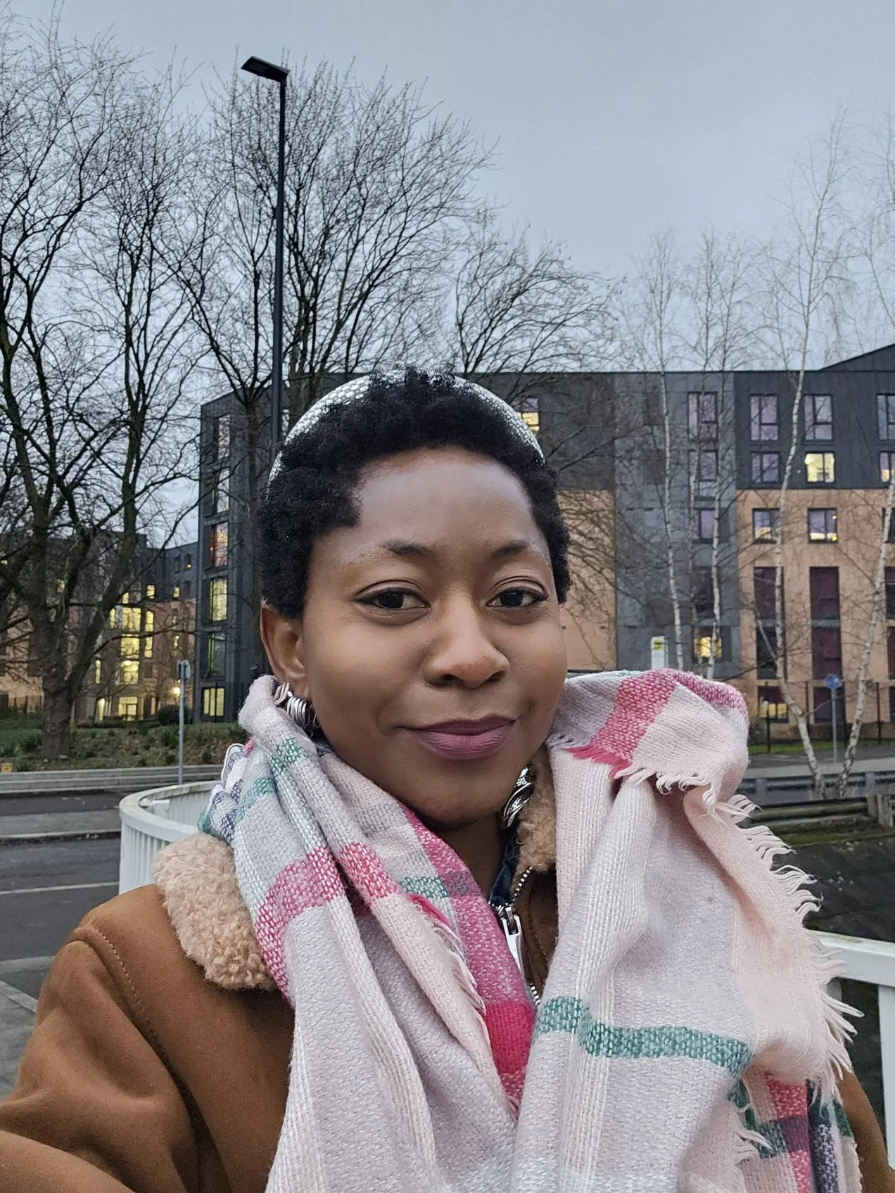 A woman taking a selfie outdoors, with leafless trees and modern apartment buildings in the background, during overcast weather.