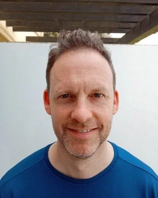 A man with short dark hair and facial hair smiling outdoors, wearing a blue shirt.
